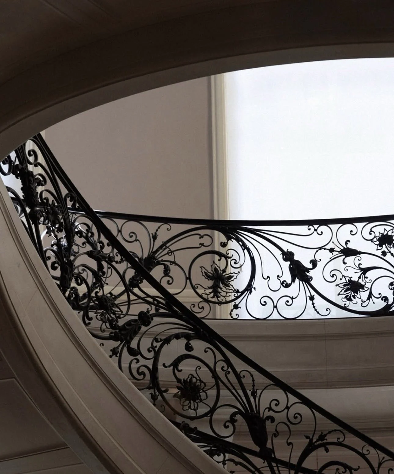 Ornate black wrought iron staircase railing with floral designs, viewed from below against a beige wall and a bright window.