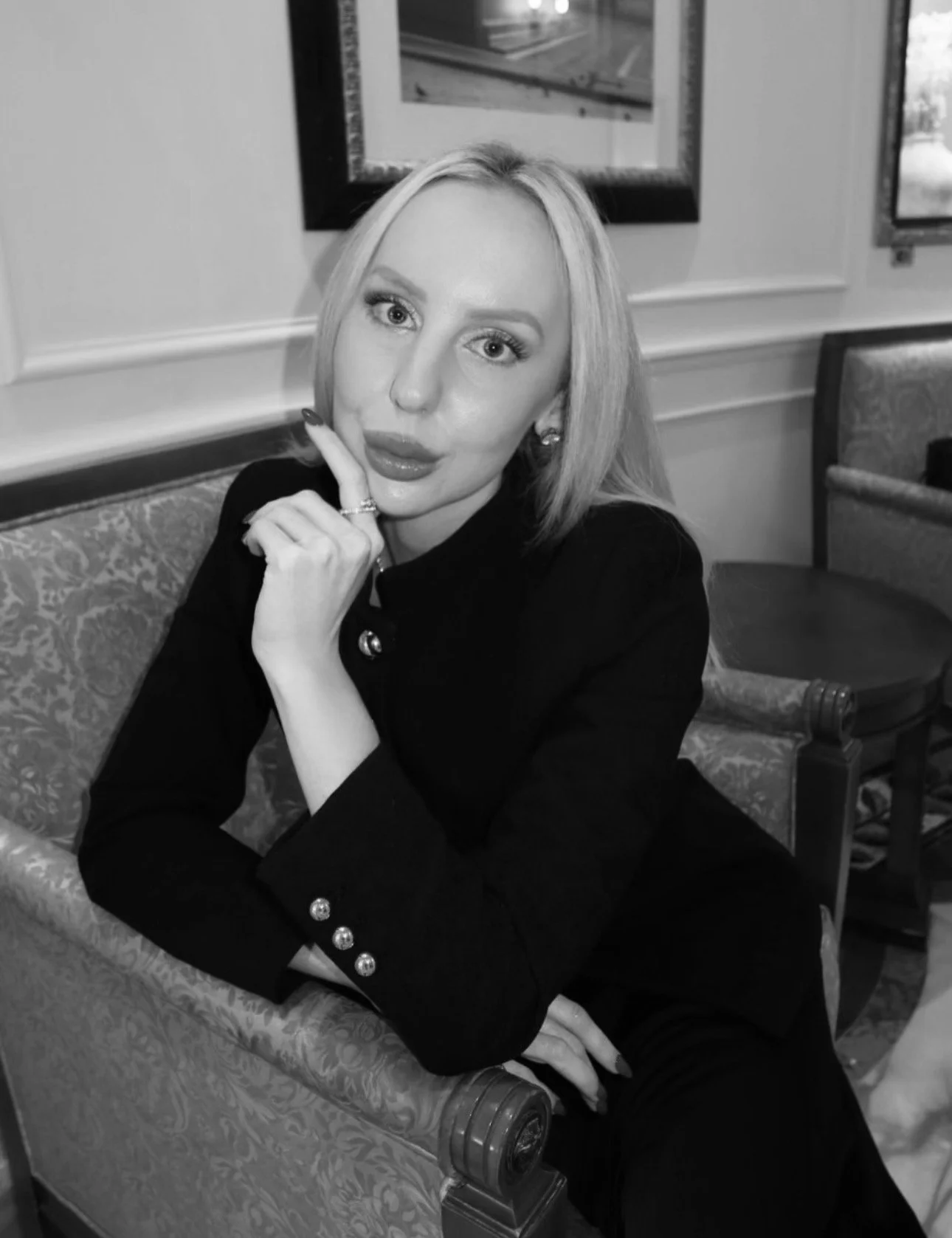 Black and white photo of a woman sitting on a floral-patterned couch with her chin resting on her hand, wearing a dark blazer with decorative buttons, looking directly at the camera.