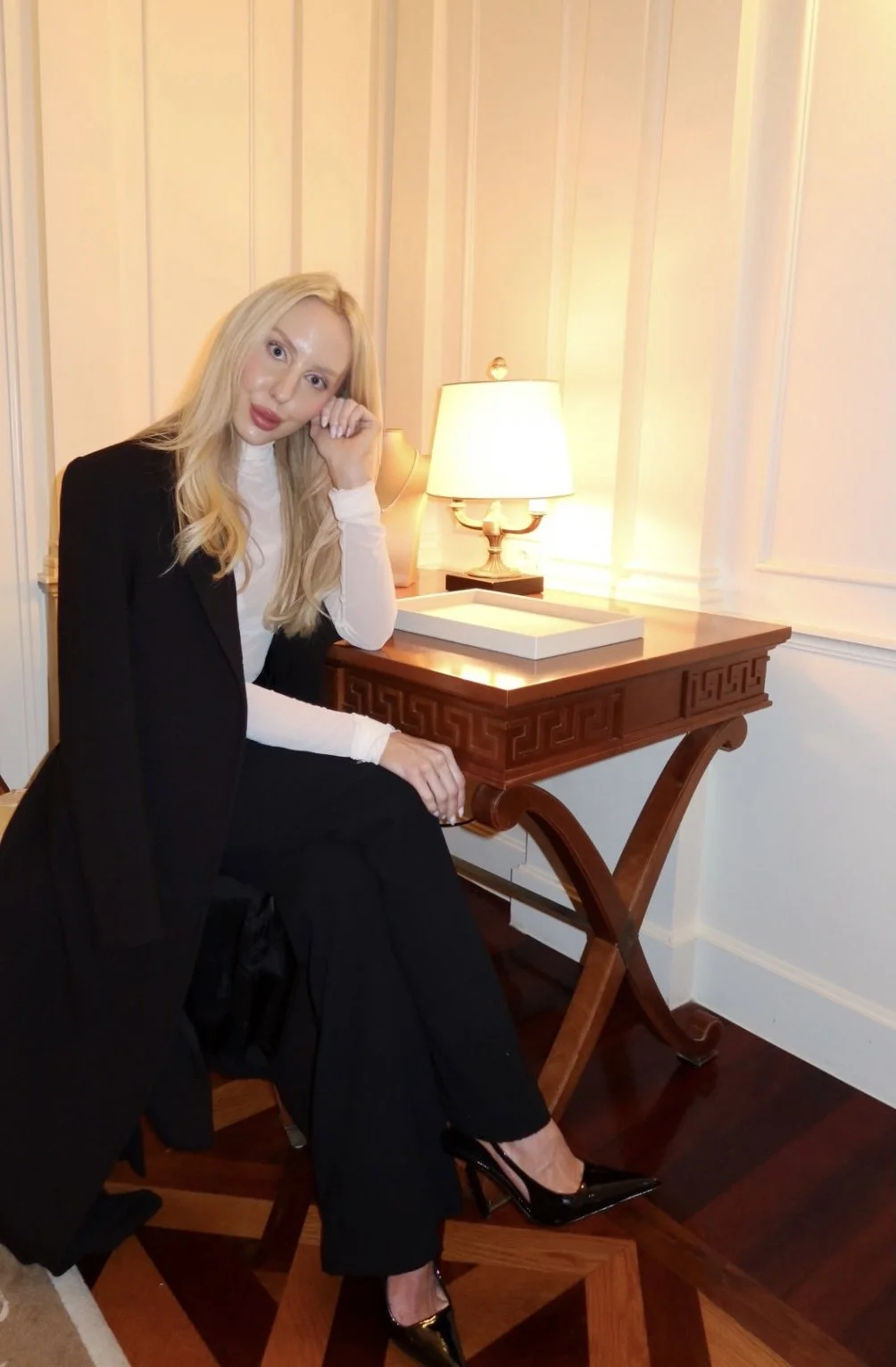 A woman with long blonde hair, wearing a black blazer, white turtleneck, black pants, and black high heels, sitting beside a wooden table with a lamp on top in a room with white-paneled walls.