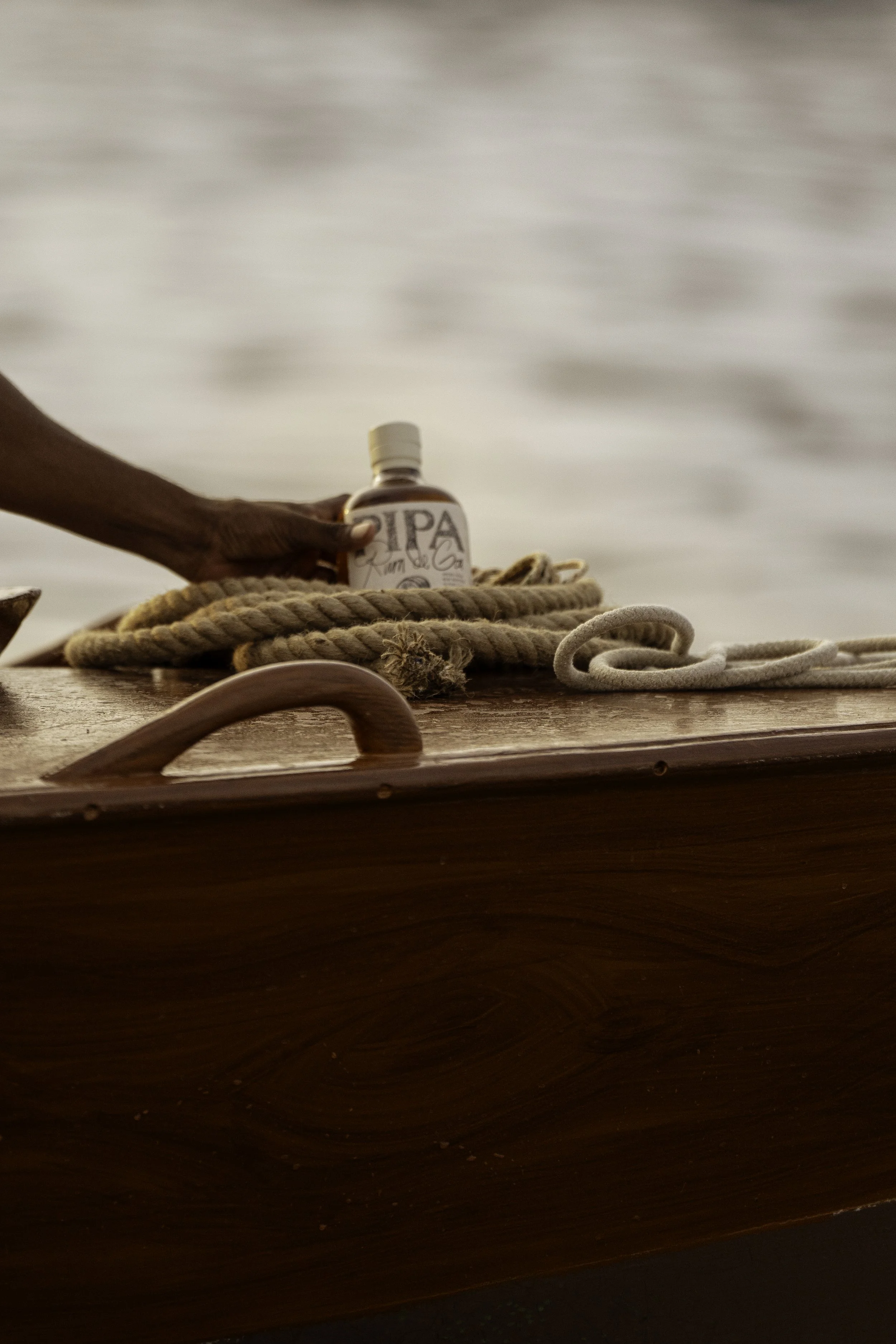 Close-up of a hand holding a small bottle labeled 'IPA' on a wooden boat, with ropes and water in the background.
