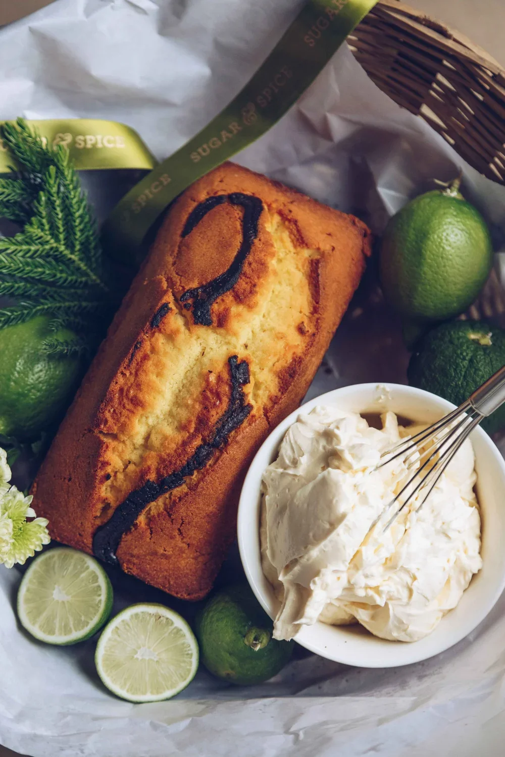 Lemon cake with a question mark drawn on top, fresh limes, a bowl of whipped cream with a whisk, and a sprig of greenery in a basket. Food Styling