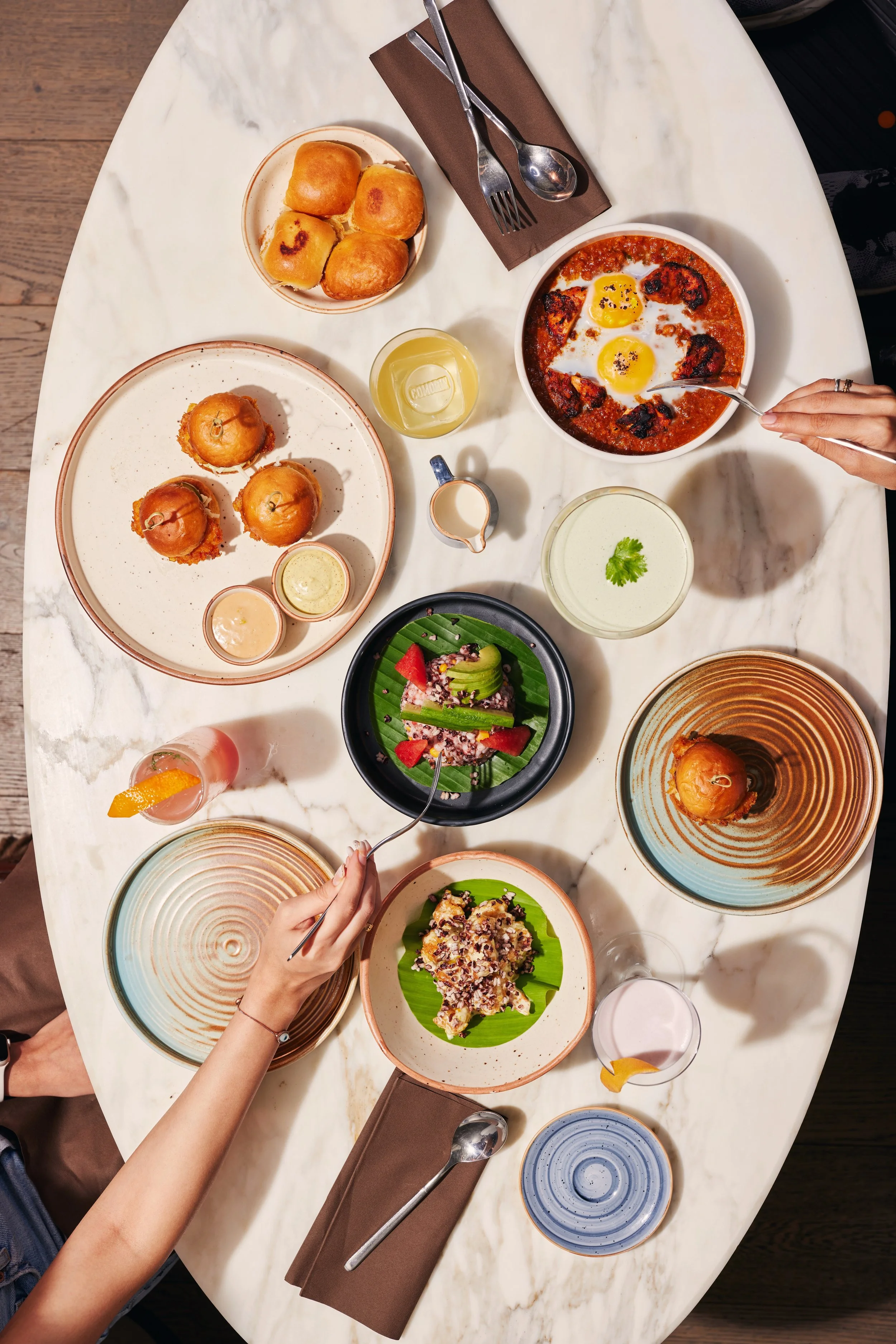A marble table with various plates of food, drinks, and cutlery. There are small bread rolls, a bowl with eggs in tomato sauce, and a bowl with salad. A person's hand is seen reaching for food, and another hand is holding a fork.