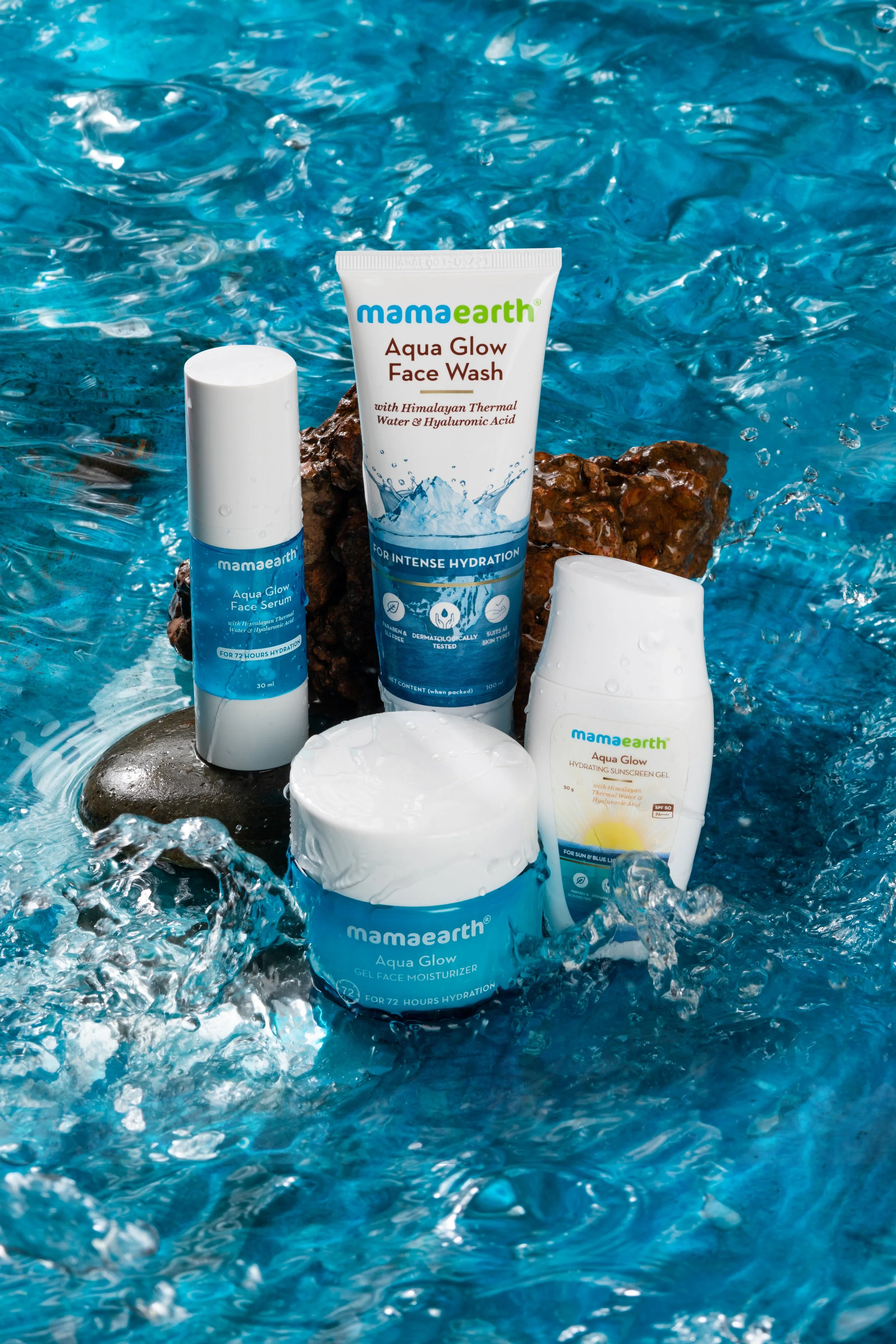 Assorted Mamaearth skincare products, including aqua glow face wash, serum, and sunscreen, arranged on rocks in a flowing water setting. Product Photography by 31-12 Studio