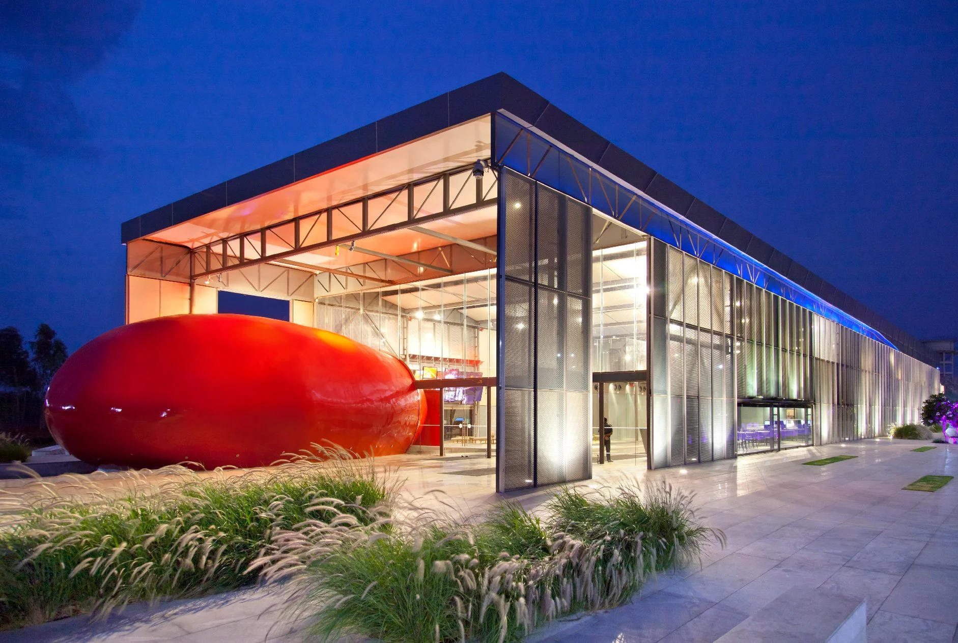 Modern building with glass walls illuminated inside, featuring a large red oval sculpture outside, surrounded by landscaped grass and plants, under a dark blue evening sky.