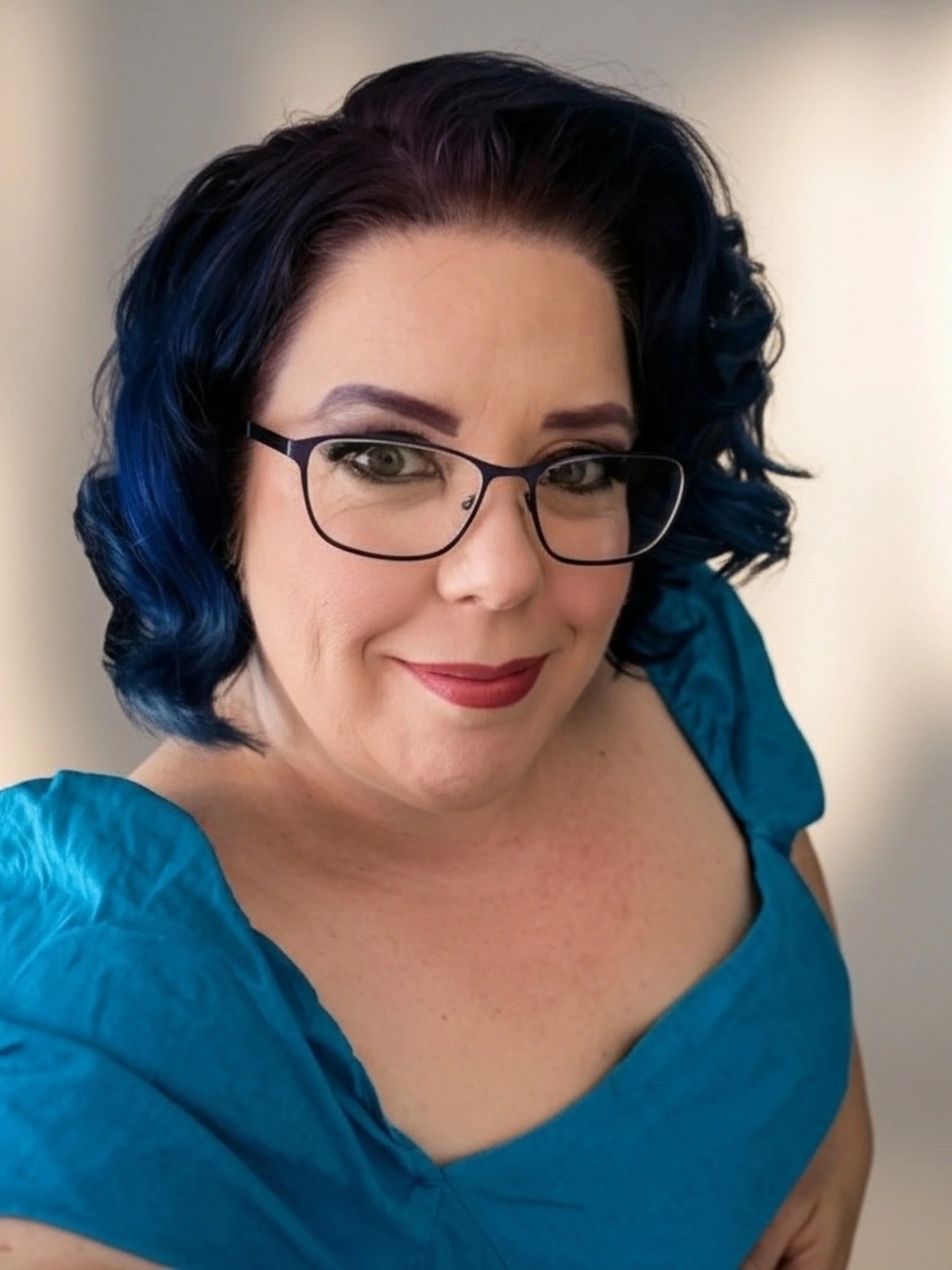 Close-up of a woman with dark curly hair with blue tips, glasses, wearing a teal blouse, smiling against a plain wall.