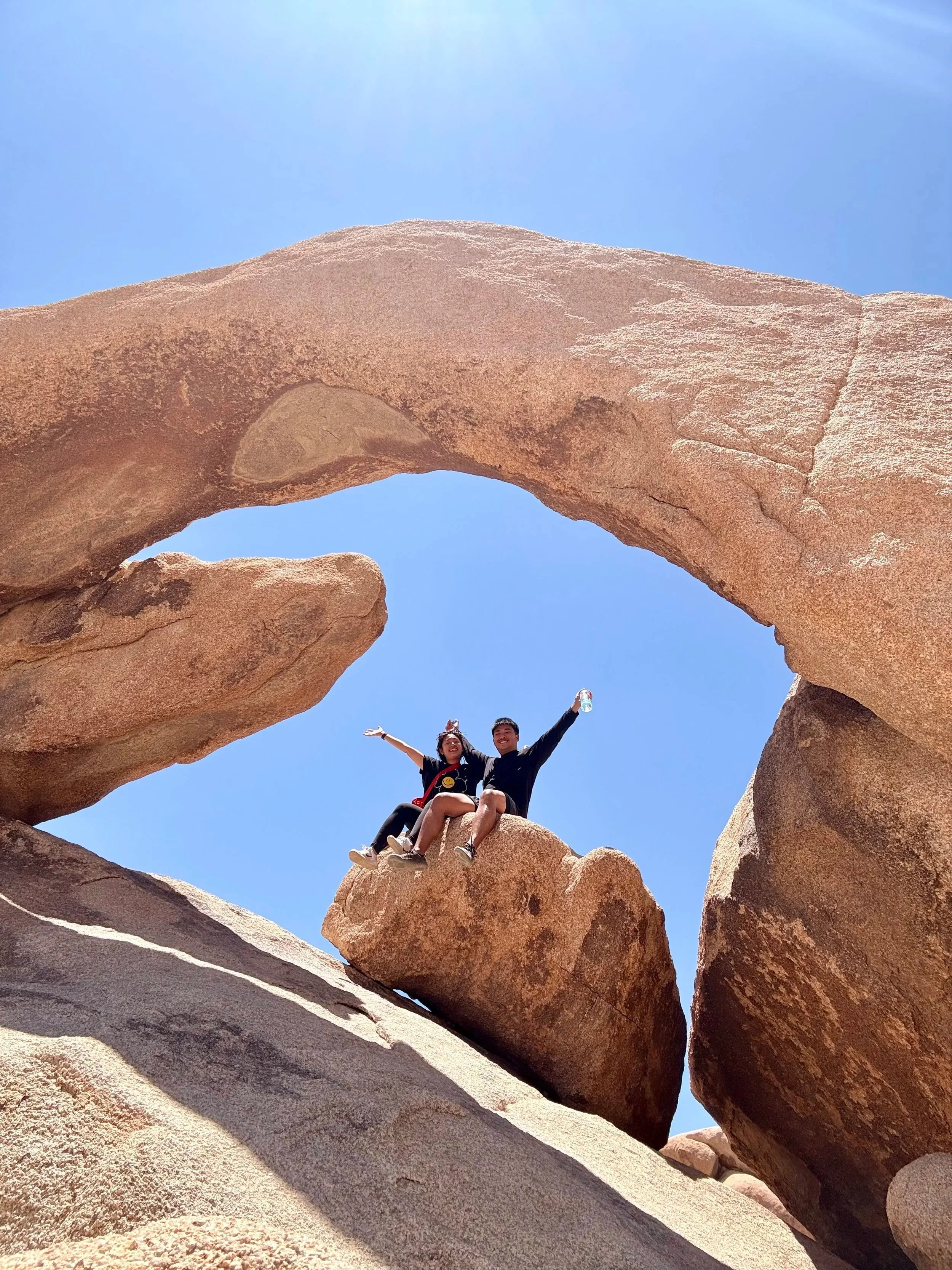 Two people are sitting on a large rock formation under a natural rock arch in a desert landscape with a clear blue sky.