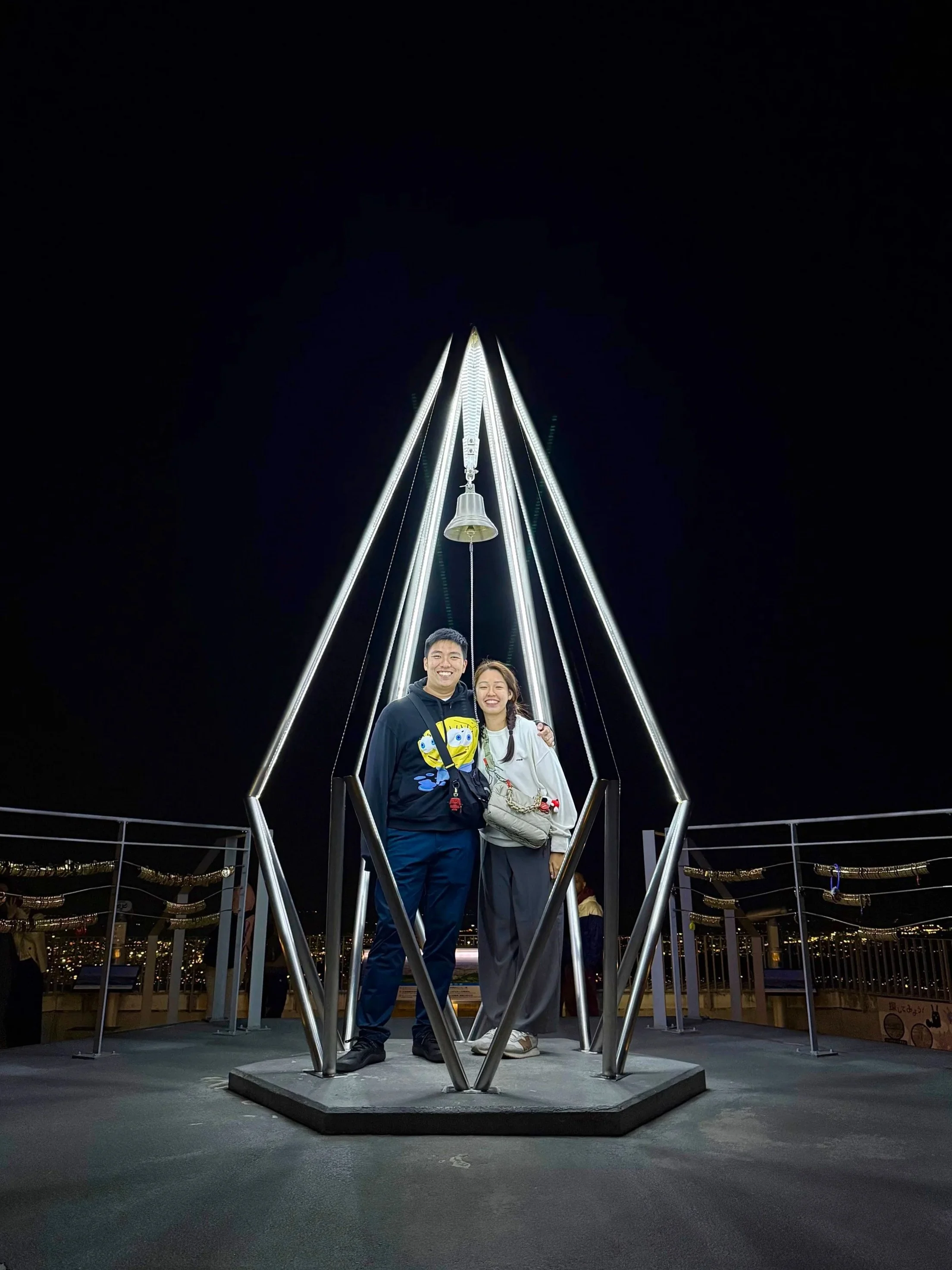 A young man and woman smiling and standing together at night on a platform with a metal bell and lighted structure above them, city lights in the background.