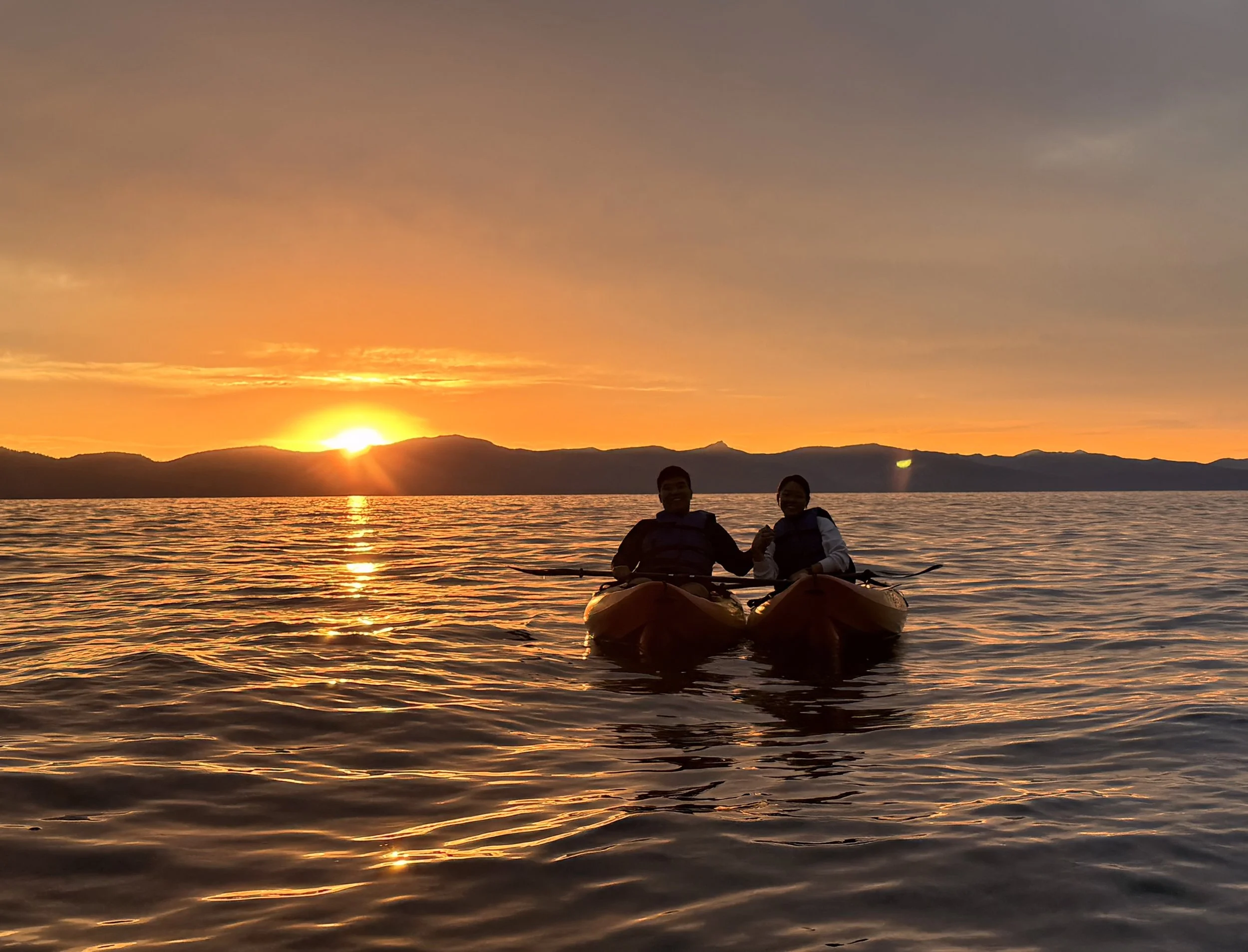 Two people kayaking on a body of water during sunset with mountains in the background.