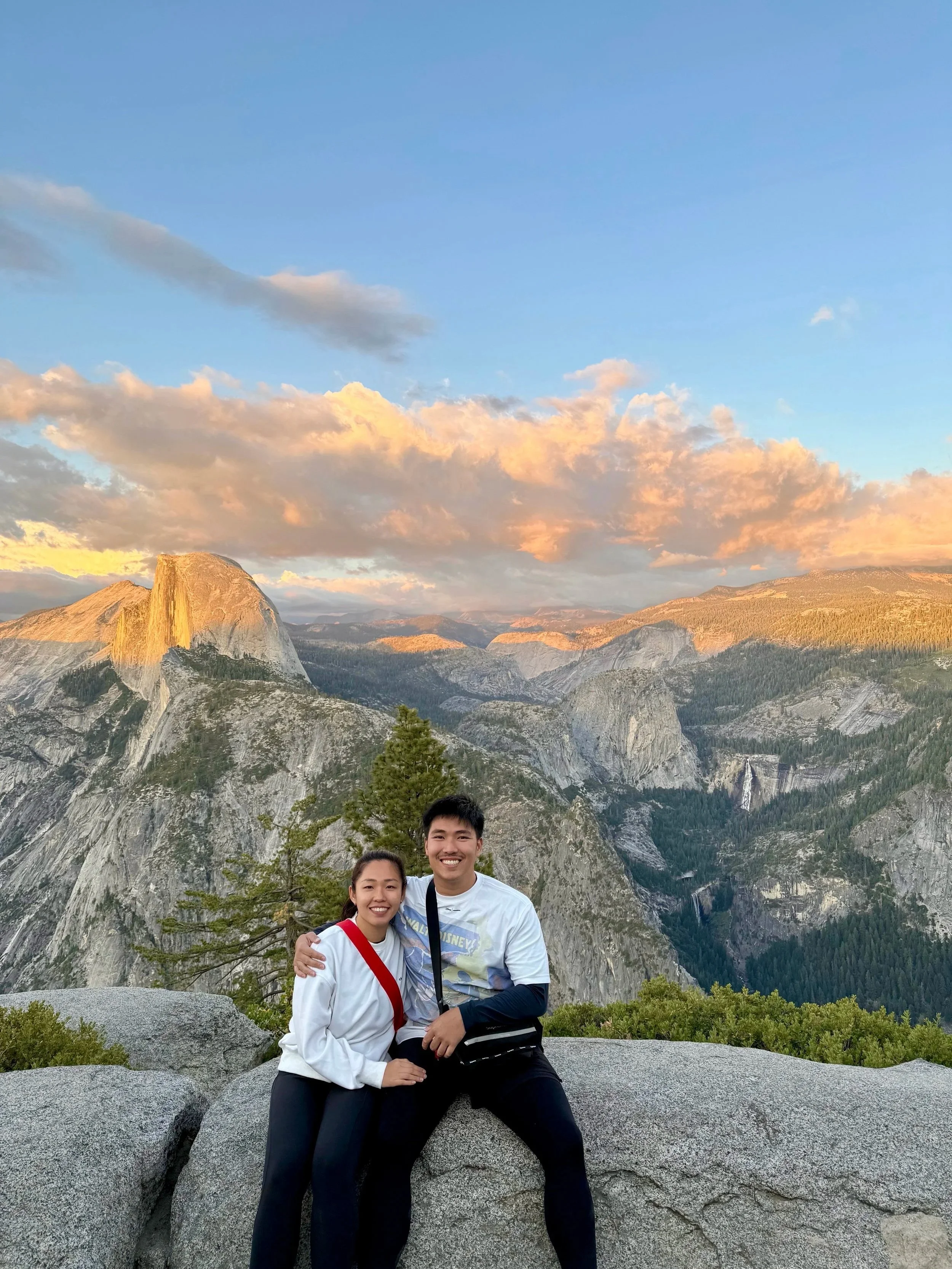 A smiling couple poses in front of a mountain landscape with clouds and a blue sky, standing on a large rock with green trees at the bottom.