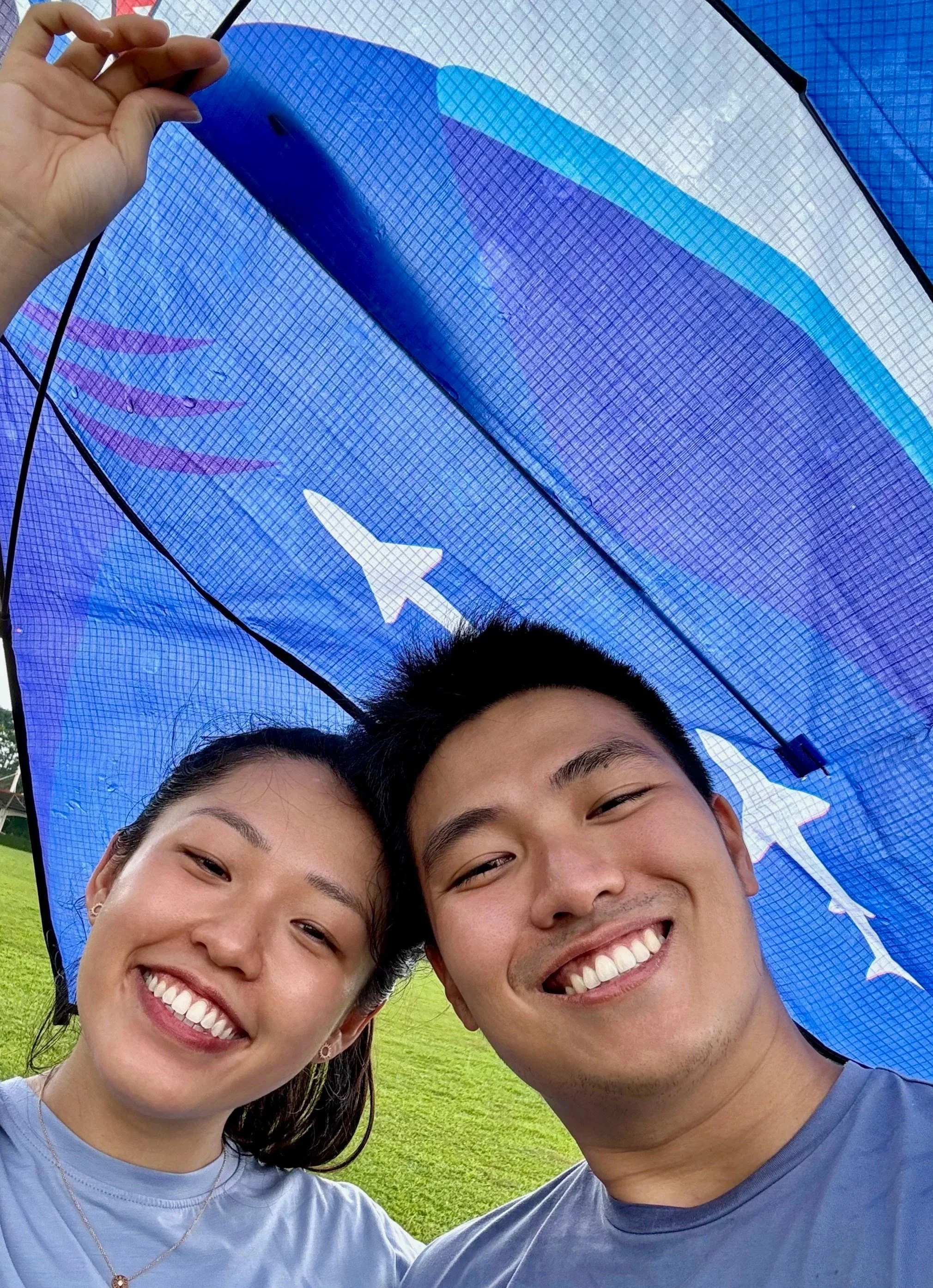 Two smiling people, a woman and a man, taking a selfie outdoors under a blue umbrella with a shark design, with a grassy field in the background.