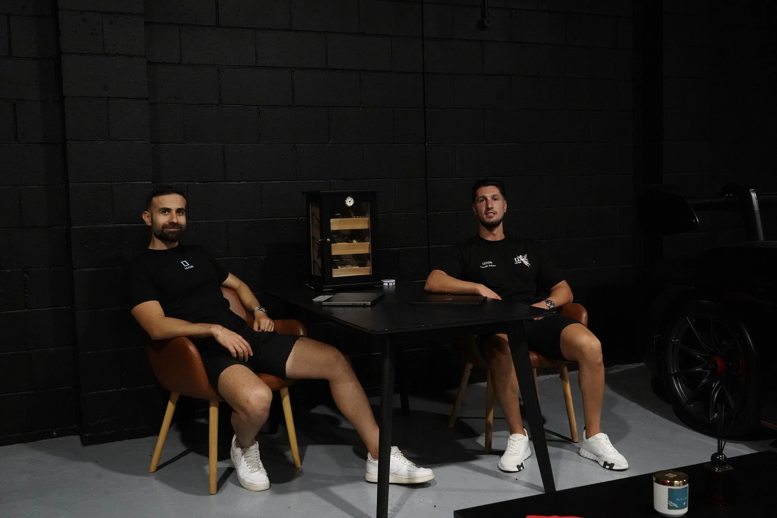 Two men seated at a table in a dark room with black brick walls, wearing black t-shirts and white sneakers, with equipment and a display case on the table.