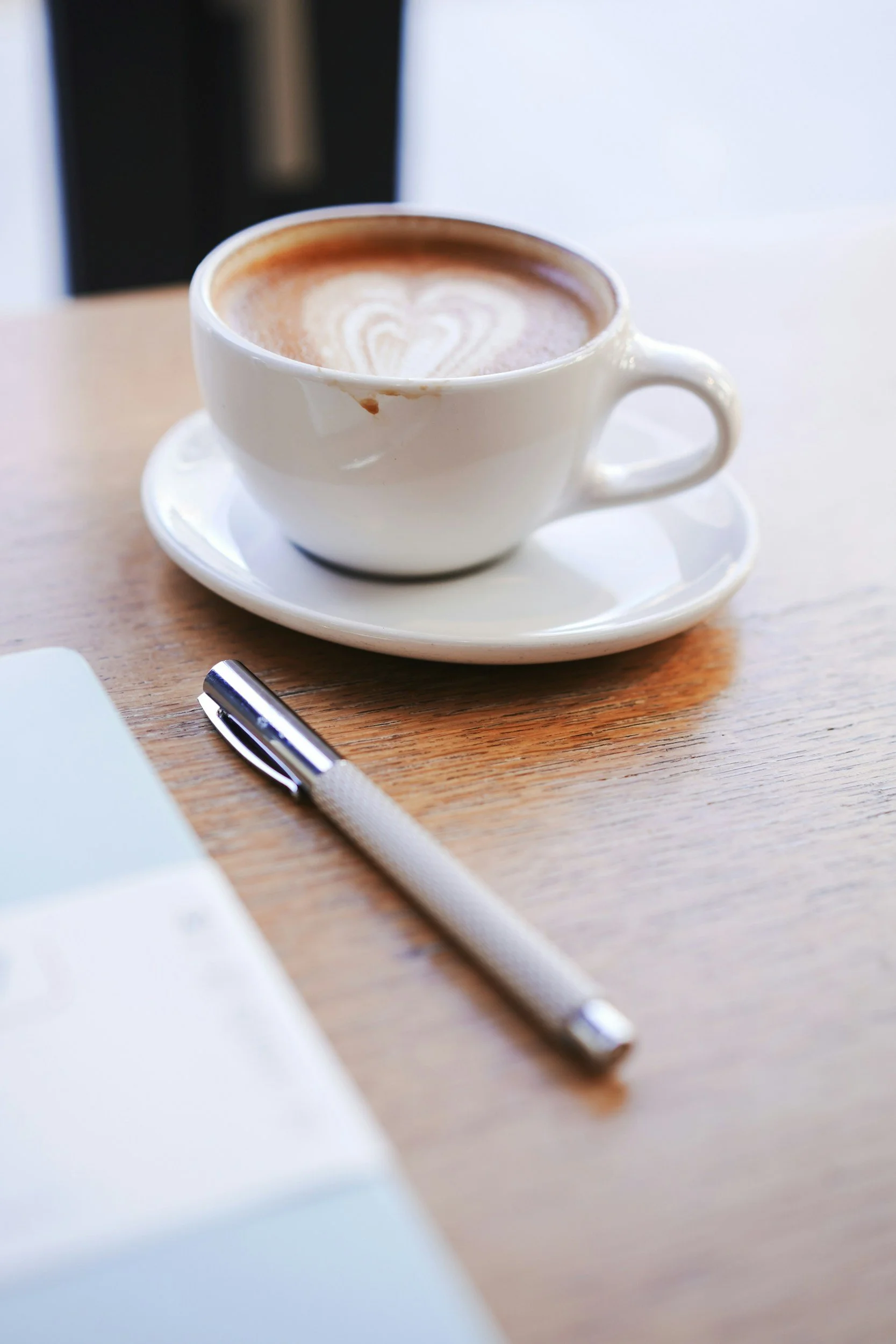 Een kop koffie met latte art op een houten tafel, naast een pen en een laptop.