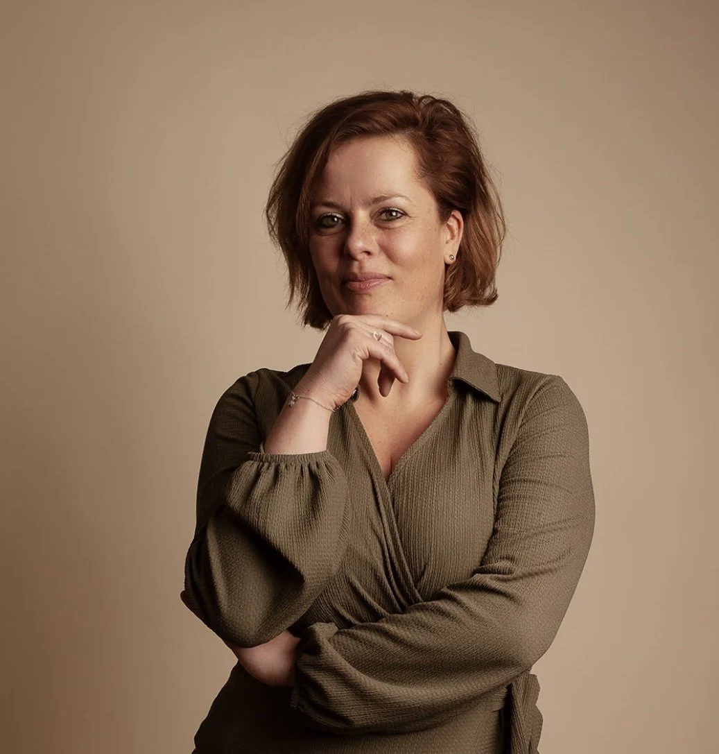 Portret van een vrouw met rood haar in een groene blouse, poseert met haar hand onder haar kin, lachend tegen een beige achtergrond.
