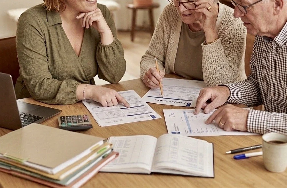 Drie ouderen zitten aan een tafel, ze bekijken financiële documenten en invullen formulieren, met een laptop, rekenmachine, en een kopje koffie op tafel.