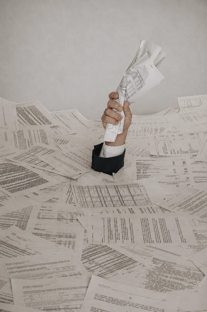 A person in a black suit jacket with a white shirt is raising a crumpled piece of paper from a desk covered with scattered documents and spreadsheets.