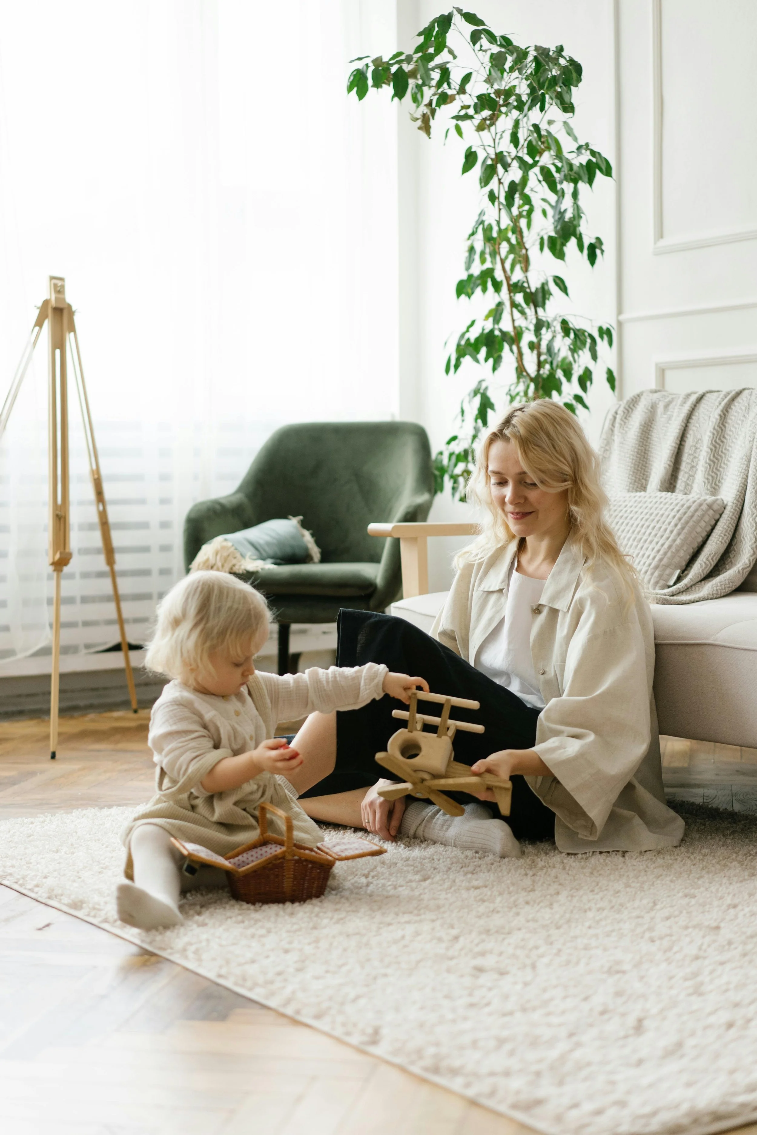 A woman with blonde hair and a young girl with blonde hair sitting on a cream rug in a warmly lit living room, playing with wooden toys. There is a basket of toys near them, a green armchair, and a tall indoor plant in the background. Soft natural light filters through sheer white curtains.