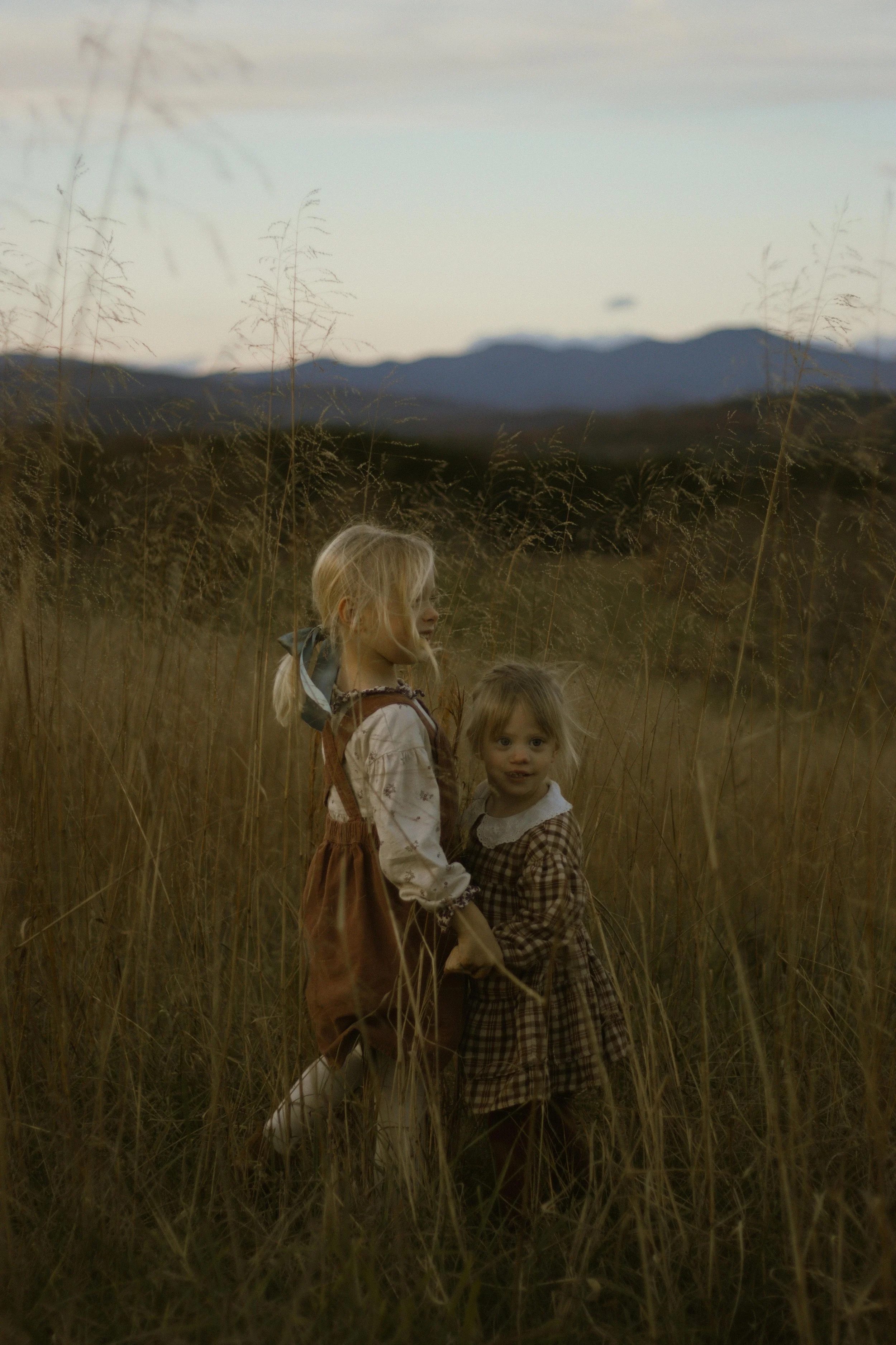 Two young children, a girl with blonde hair and a boy with light brown hair, stand in a field of tall grass with mountains in the background during sunset or dusk.
