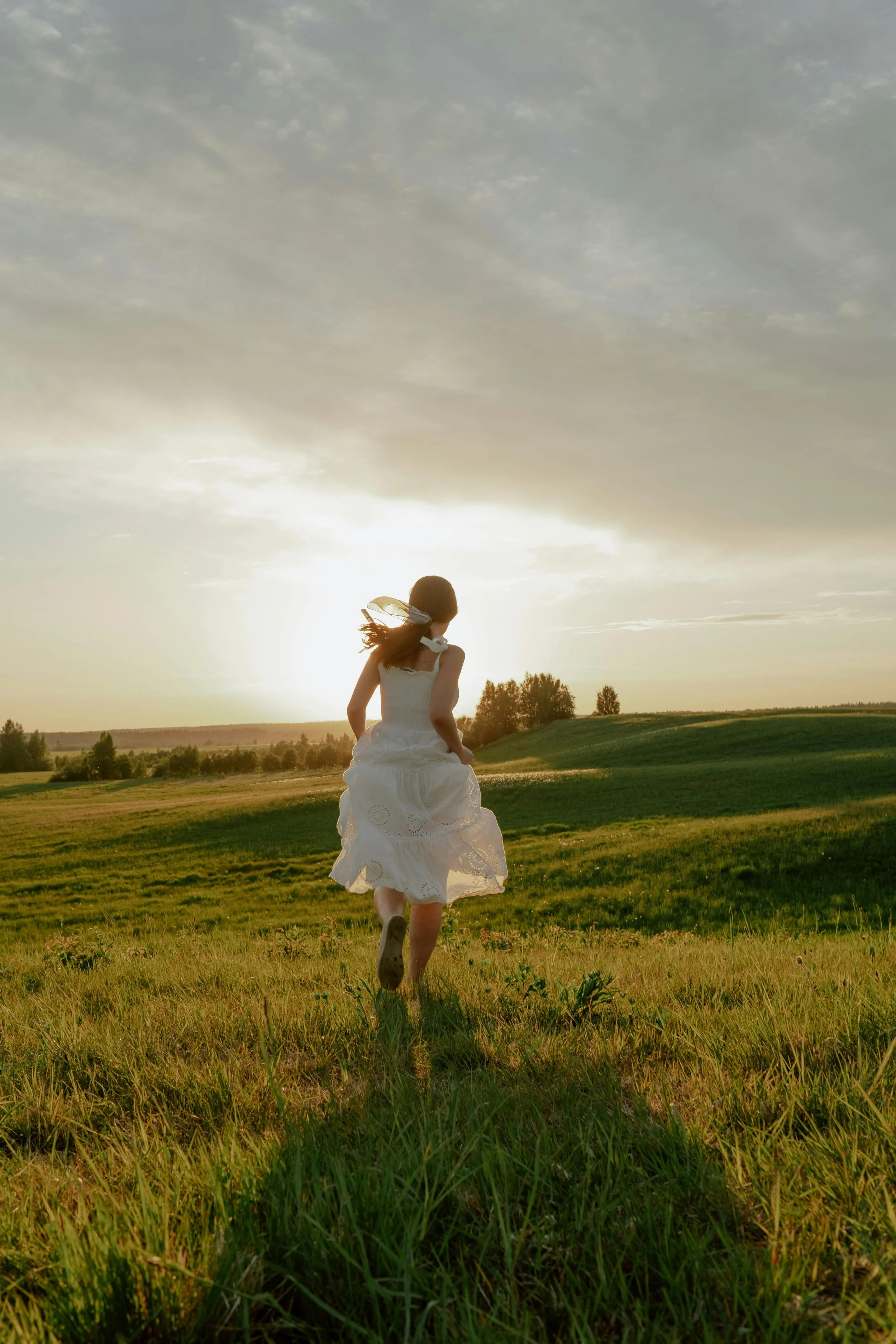 A girl in a white dress running through a grassy field during sunset or sunrise, with clouds in the sky and trees in the background.