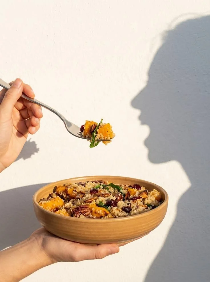 Person holding a bowl of grain salad with fruits and greens, while another person is bringing a forkful to their mouth, shadowed against a white wall.