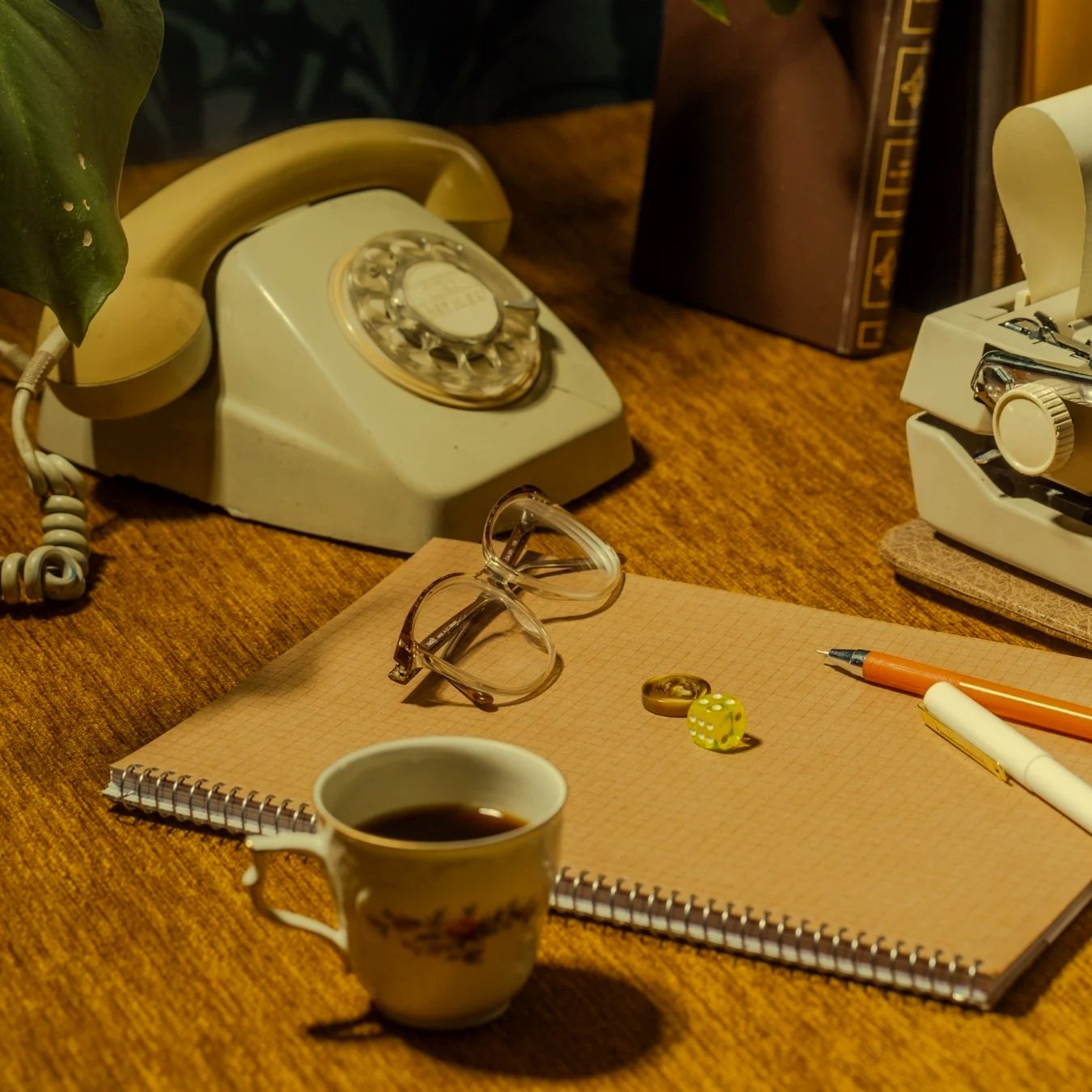 A vintage desk setup with a rotary phone, a pair of glasses, a yellow dice, a coffee cup, a notepad, a pen, a fountain pen, a typewriter, and some books on a wooden surface.