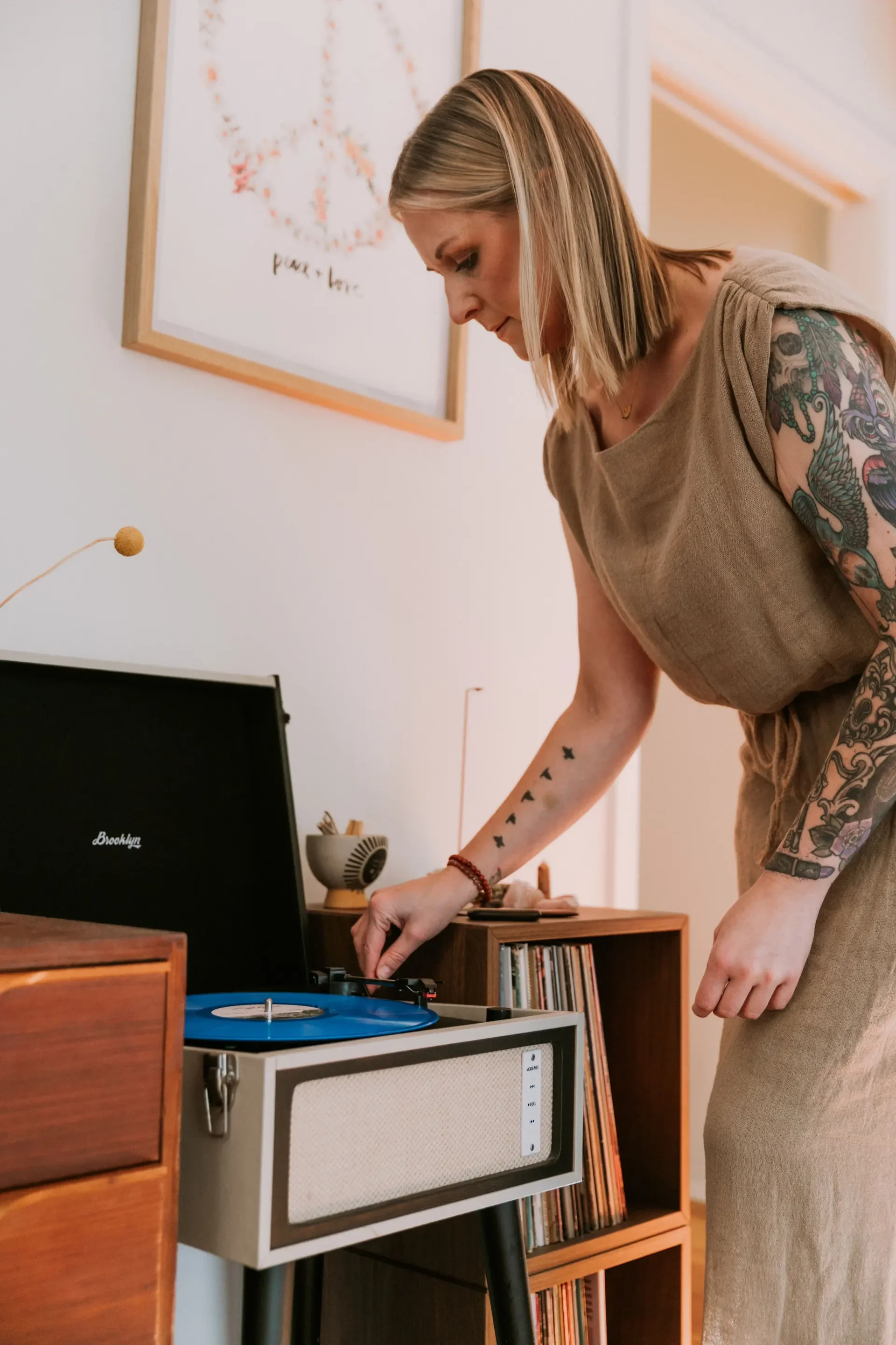 Marisa Robinson operating a vintage flat-top turntable with a blue vinyl record. The room features a wooden cabinet with vinyl records and a bookshelf. A framed sewing pattern is on the wall.