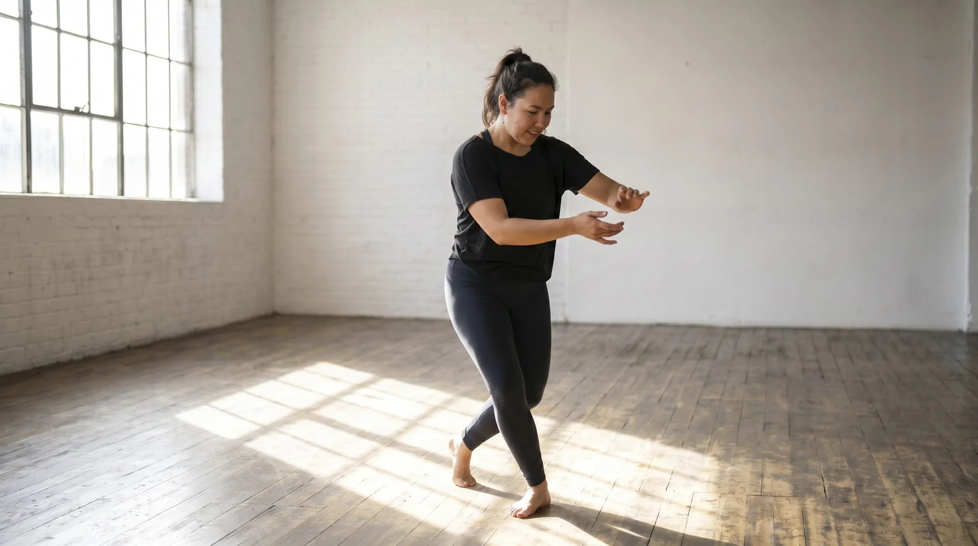 A woman in black athletic wear practicing a dance move or yoga pose in a bright, spacious studio with large windows and wooden floor.