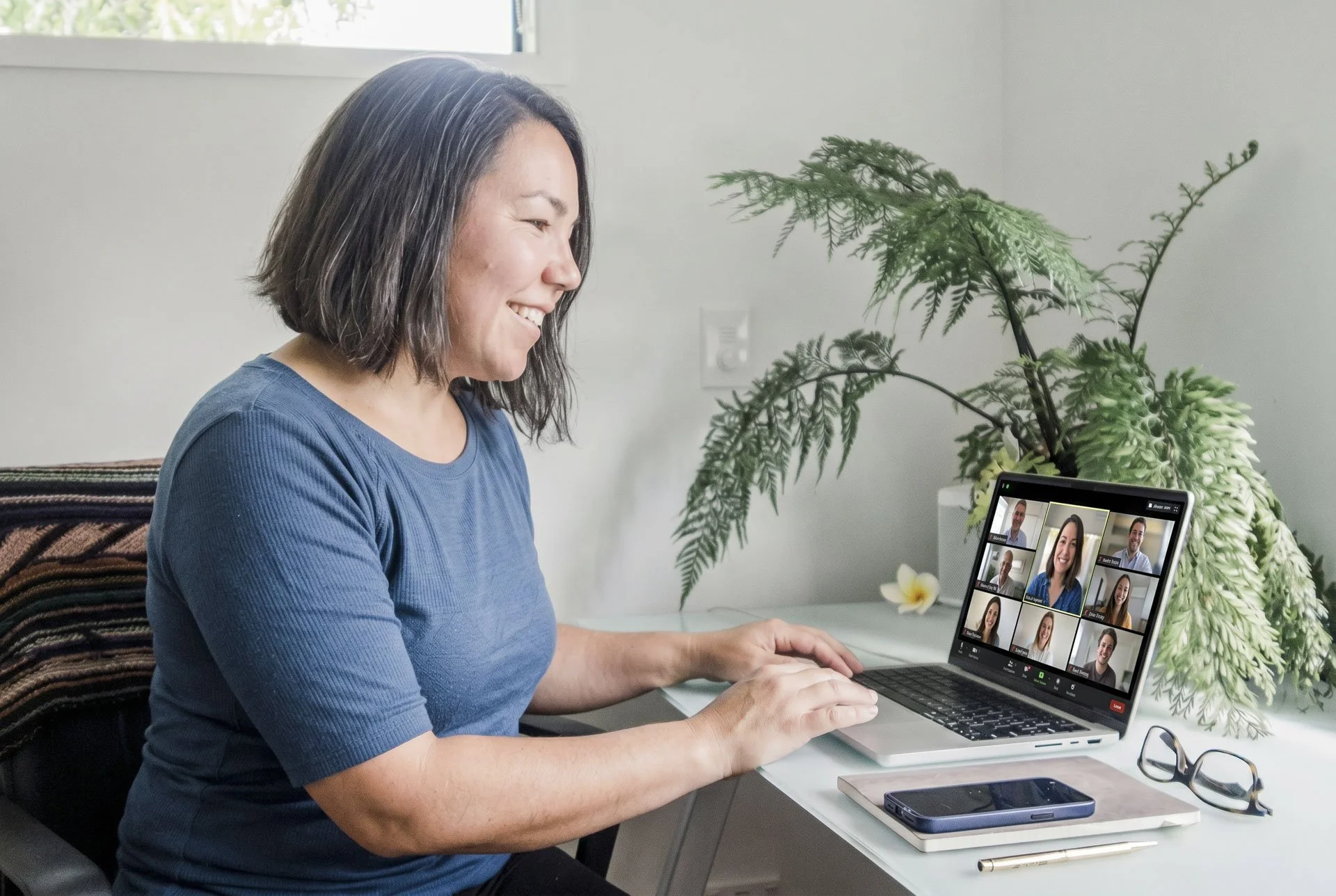 Woman participating in a video conference on her laptop, sitting at a desk with a plant in the background.