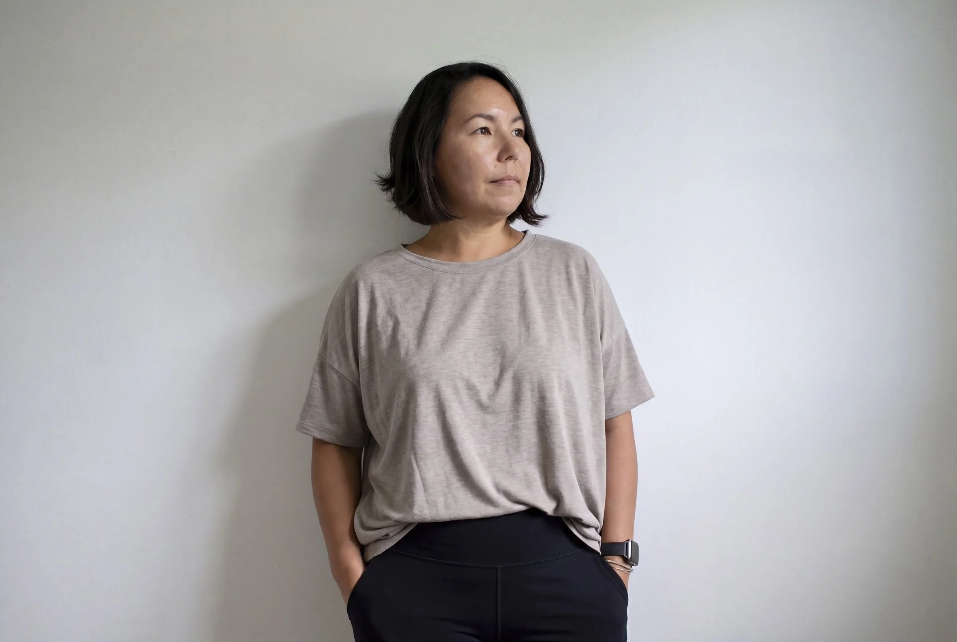 A woman with shoulder-length dark hair standing against a plain white wall, wearing a loose beige t-shirt, black pants, a watch on her wrist, and looking off to the side.