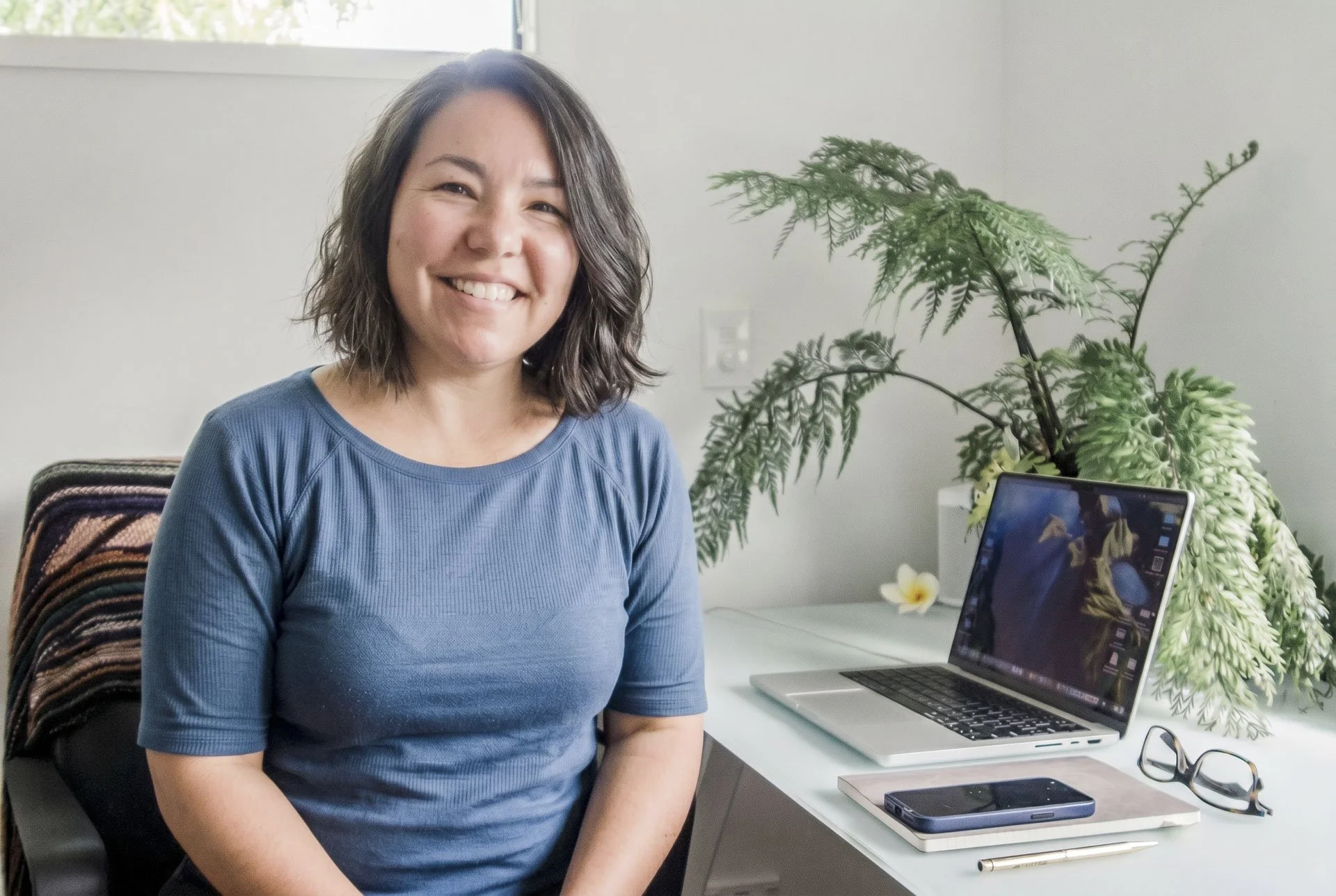 A woman with shoulder-length dark hair, smiling and wearing a blue shirt, sits at a white desk with a laptop, smartphone, and glasses, in a room with a large green plant and a window in the background.