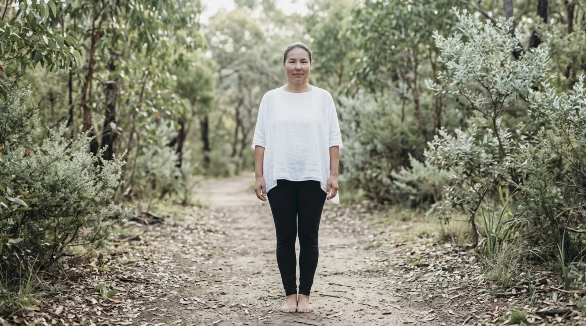 A woman stands barefoot on a dirt trail in a green forest, wearing a white top and black pants, with trees and foliage in the background.