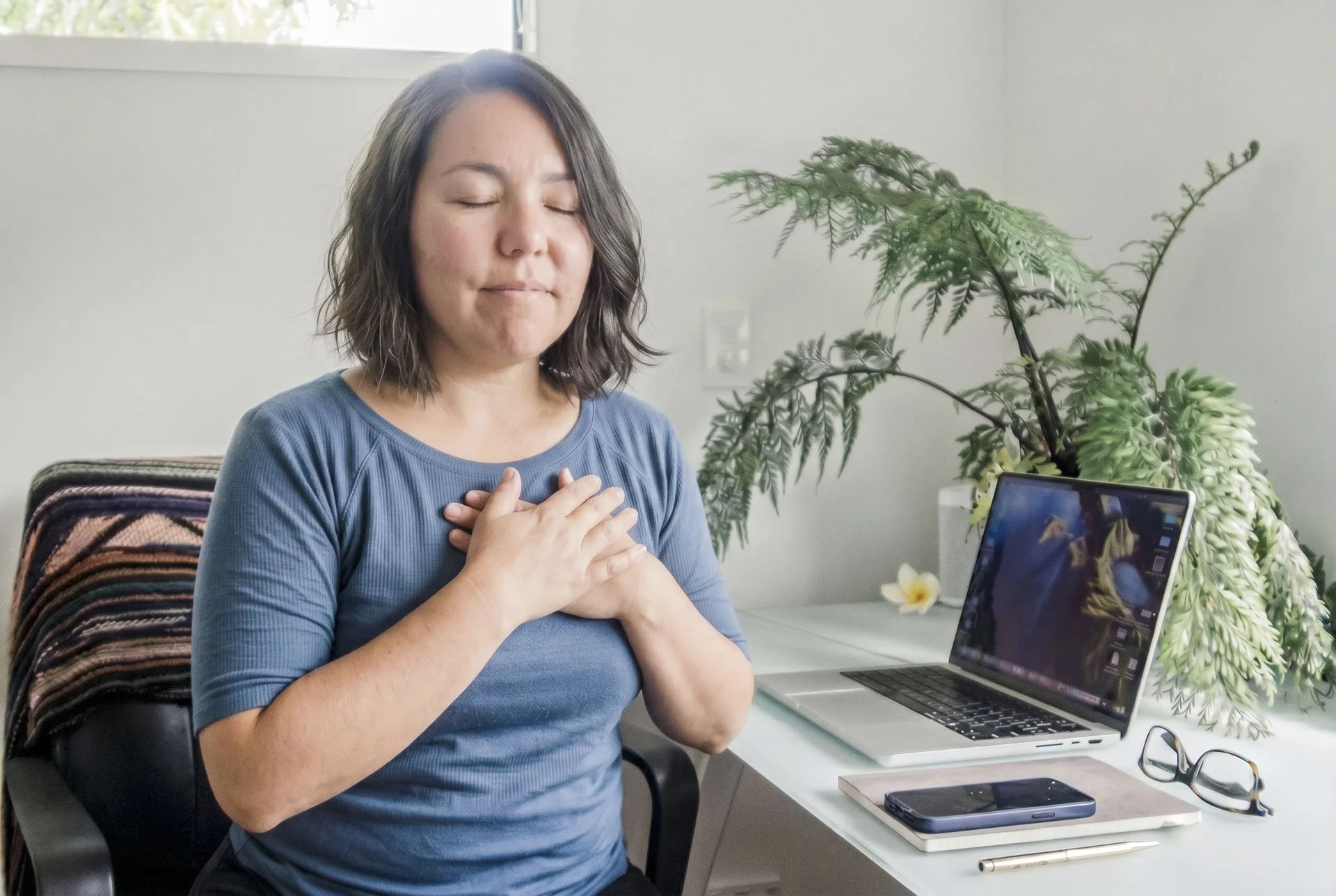 A woman with shoulder-length dark hair and a blue shirt sits at a white desk with her hands on her chest, eyes closed, and a peaceful expression. There is a large green leafy plant, a laptop, a smartphone, a closed notebook, and a pair of glasses on the desk. Behind her is a window and a neutral wall.