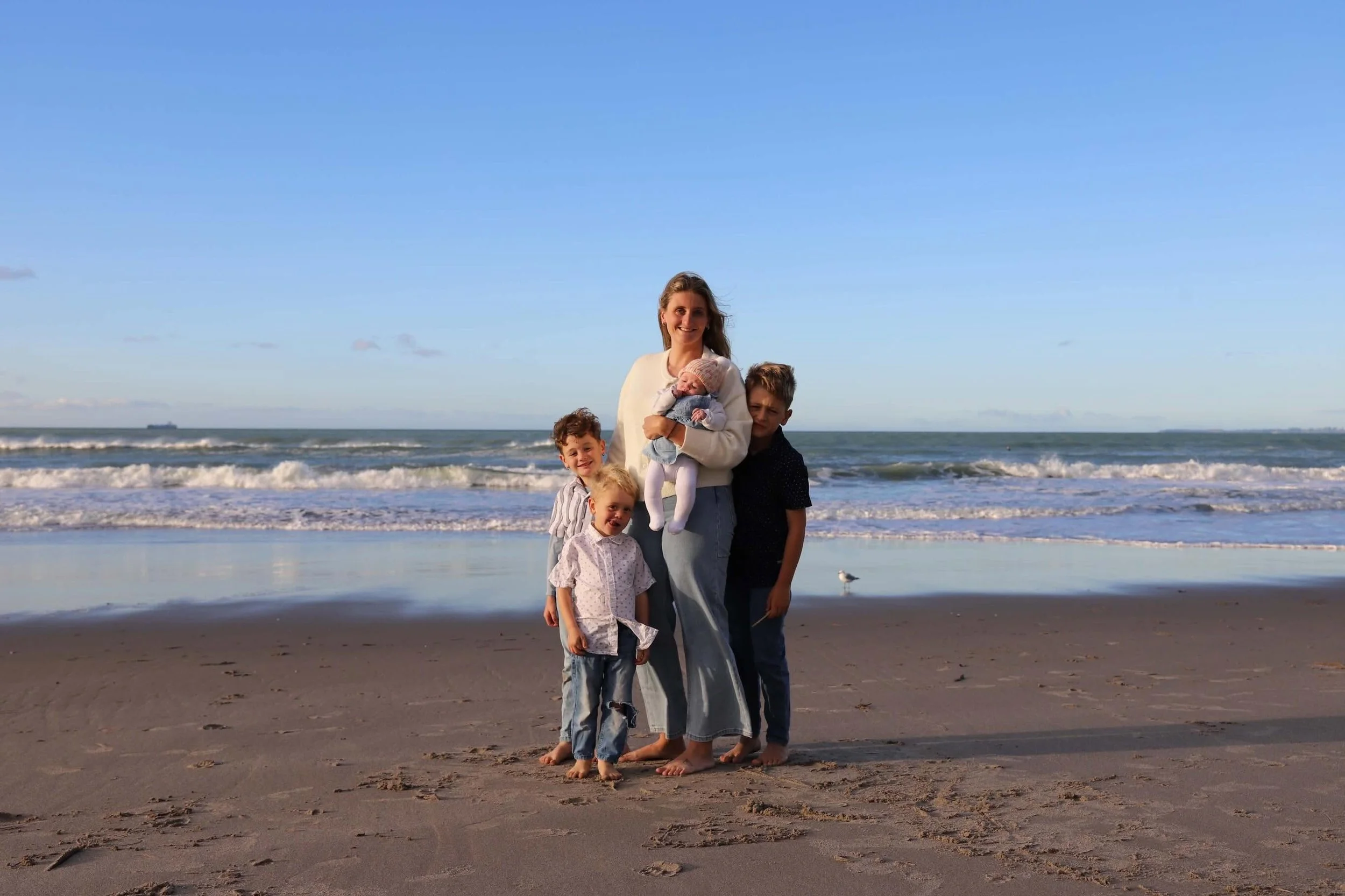 A woman and four children standing on a sandy beach with the ocean and blue sky in the background. The woman is holding a baby, and the children are smiling.