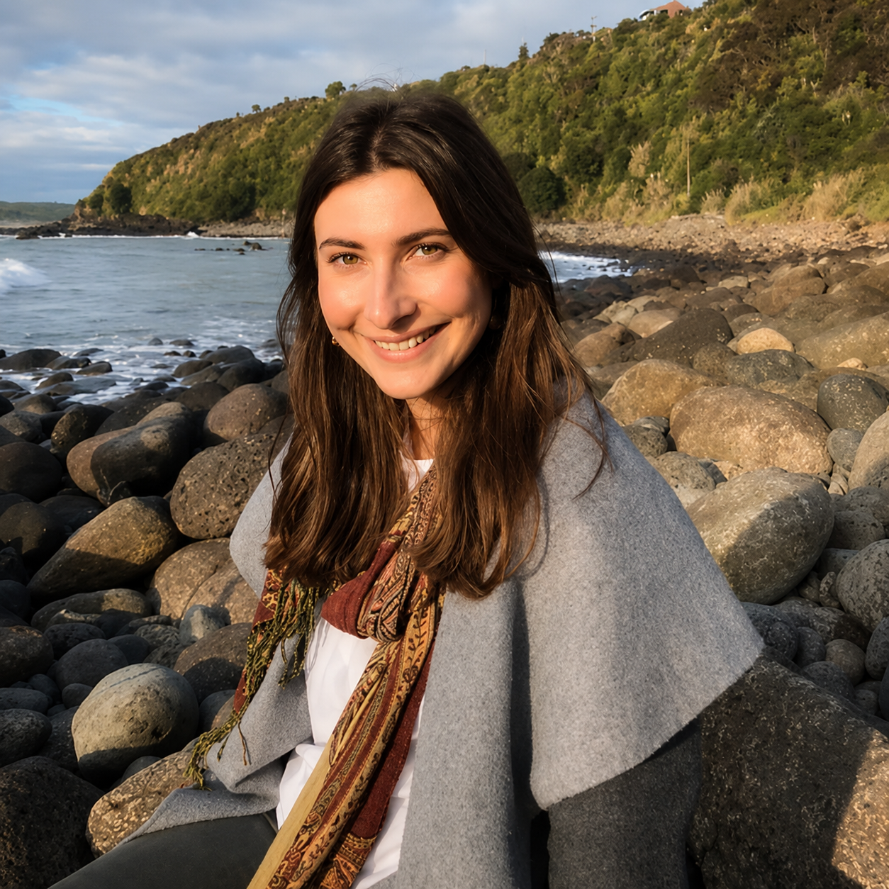 A smiling woman with long brown hair sitting on rocks at a beach with water, a hillside, and a cloudy sky in the background during sunset.