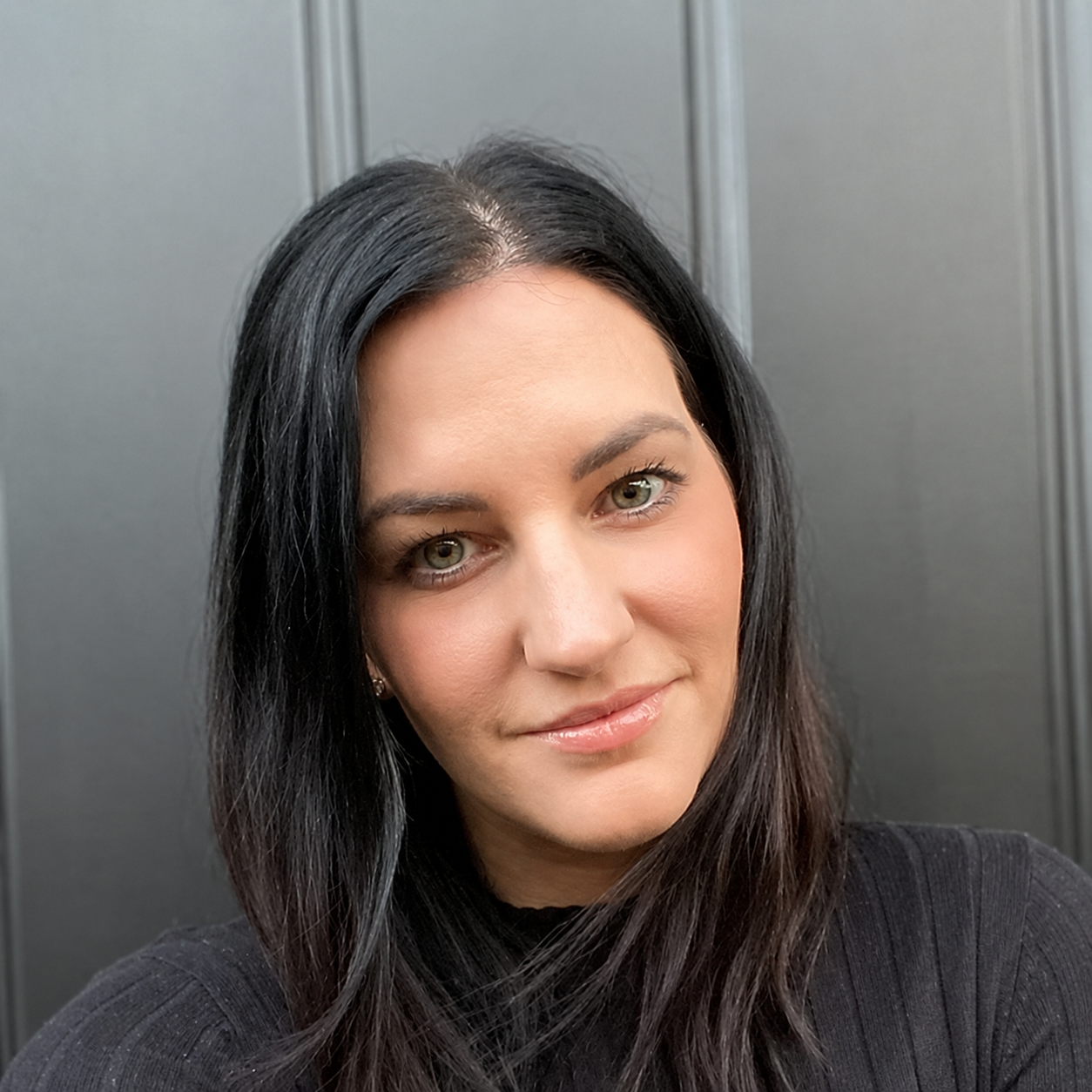 Close-up of a woman with long dark hair, light skin, and brown eyes, smiling softly, wearing a black top, standing against a gray background.
