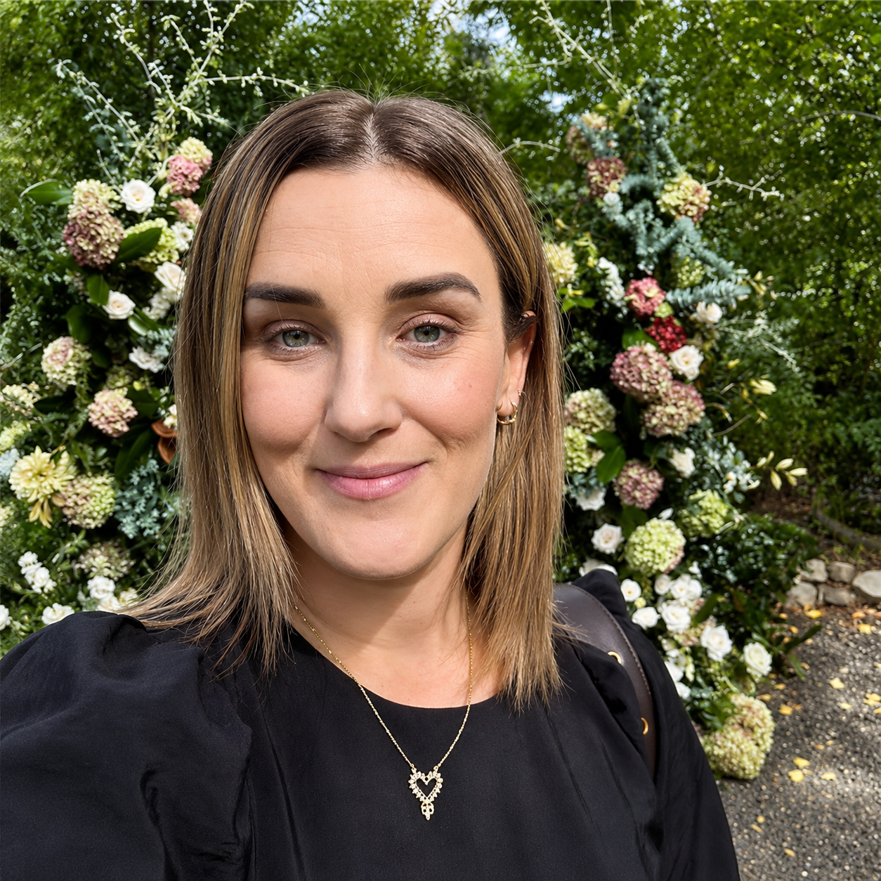 A woman with shoulder-length light brown hair, wearing a black top and heart-shaped pendant necklace, standing outdoors in front of a floral arrangement with various white, pink, and green flowers and foliage.