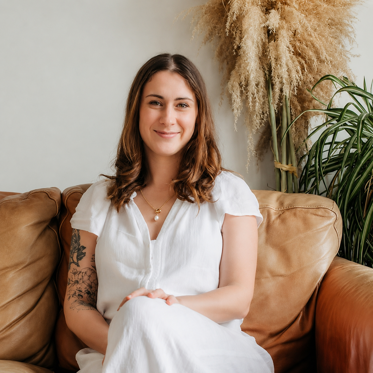 A young woman with brown wavy hair, wearing a white dress and jewelry, sitting on a tan leather couch with plants and dried pampas grass in the background.