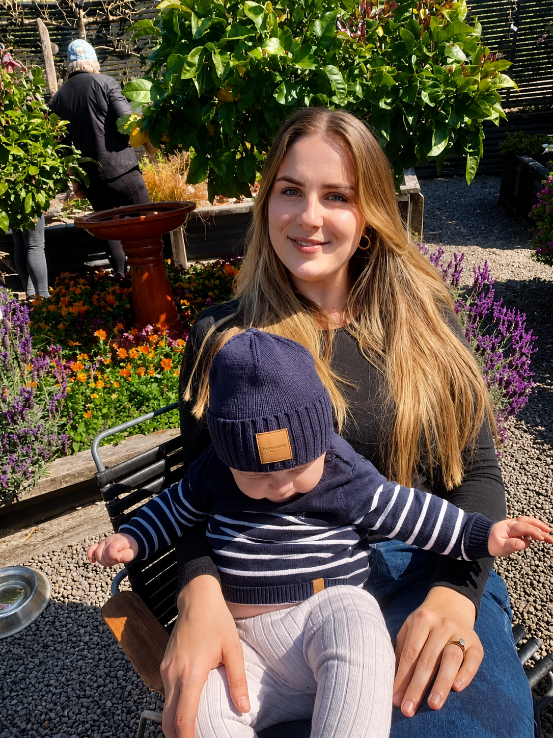 A woman with long blonde hair sitting outdoors on a bench, holding a young child on her lap. The woman is smiling, and the child is wearing a navy beanie and striped shirt, looking downward. They are in a garden with colorful flowers and lush greenery.