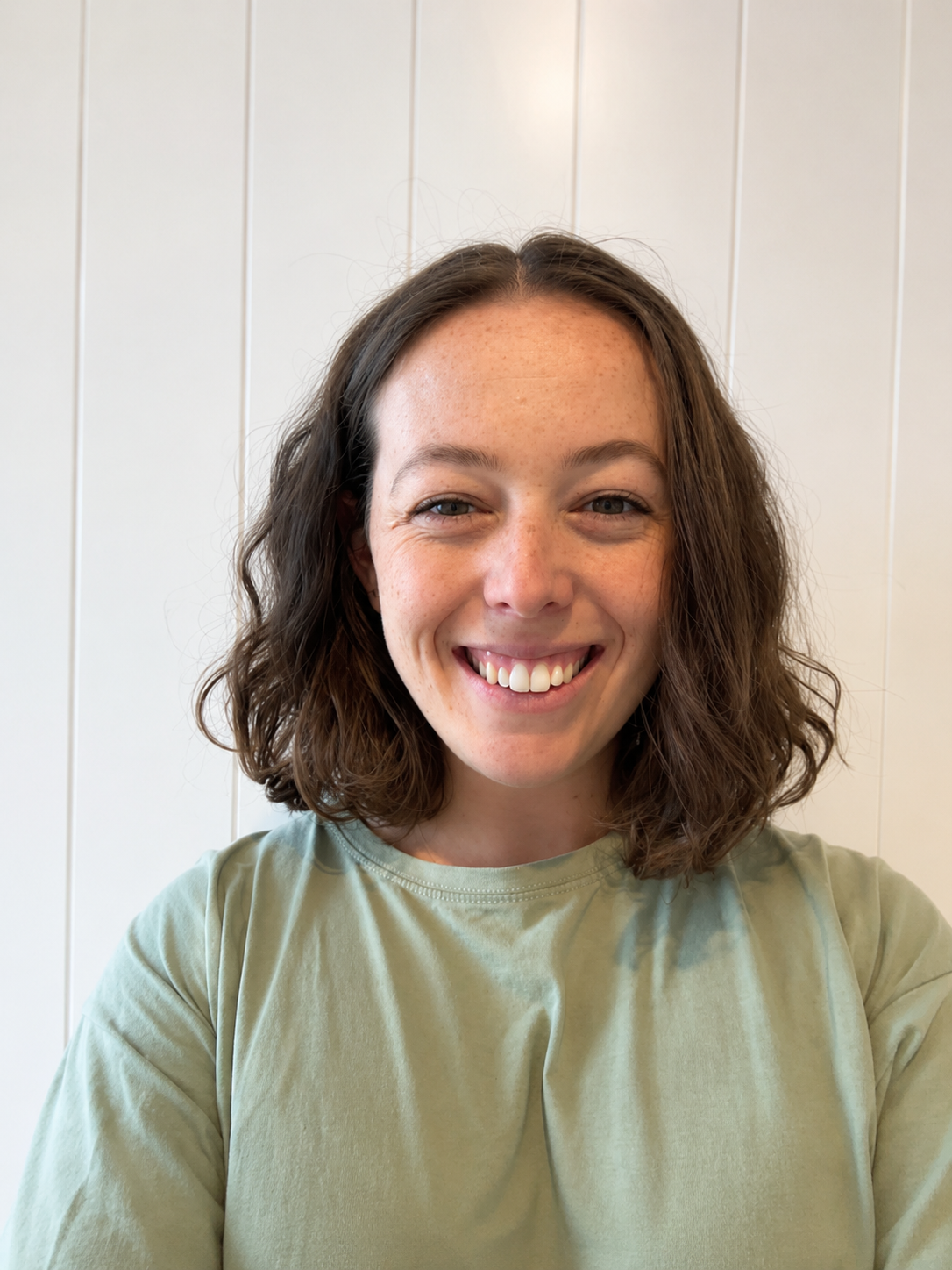 A young woman with shoulder-length curly brown hair and freckles, smiling and wearing a light green shirt, standing in front of a white paneled wall.