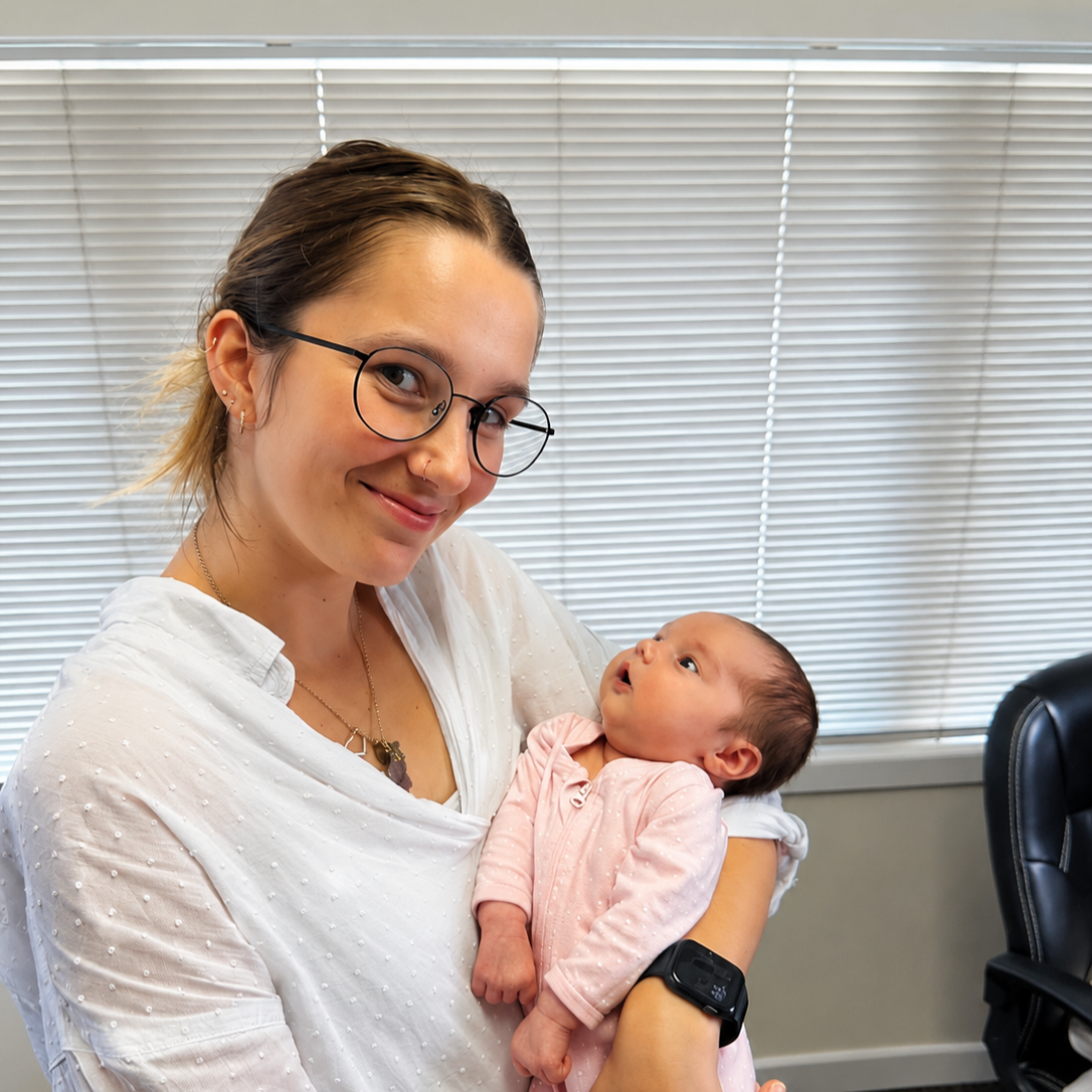 A woman with glasses holding a baby girl in a pink outfit indoors, smiling at the camera.