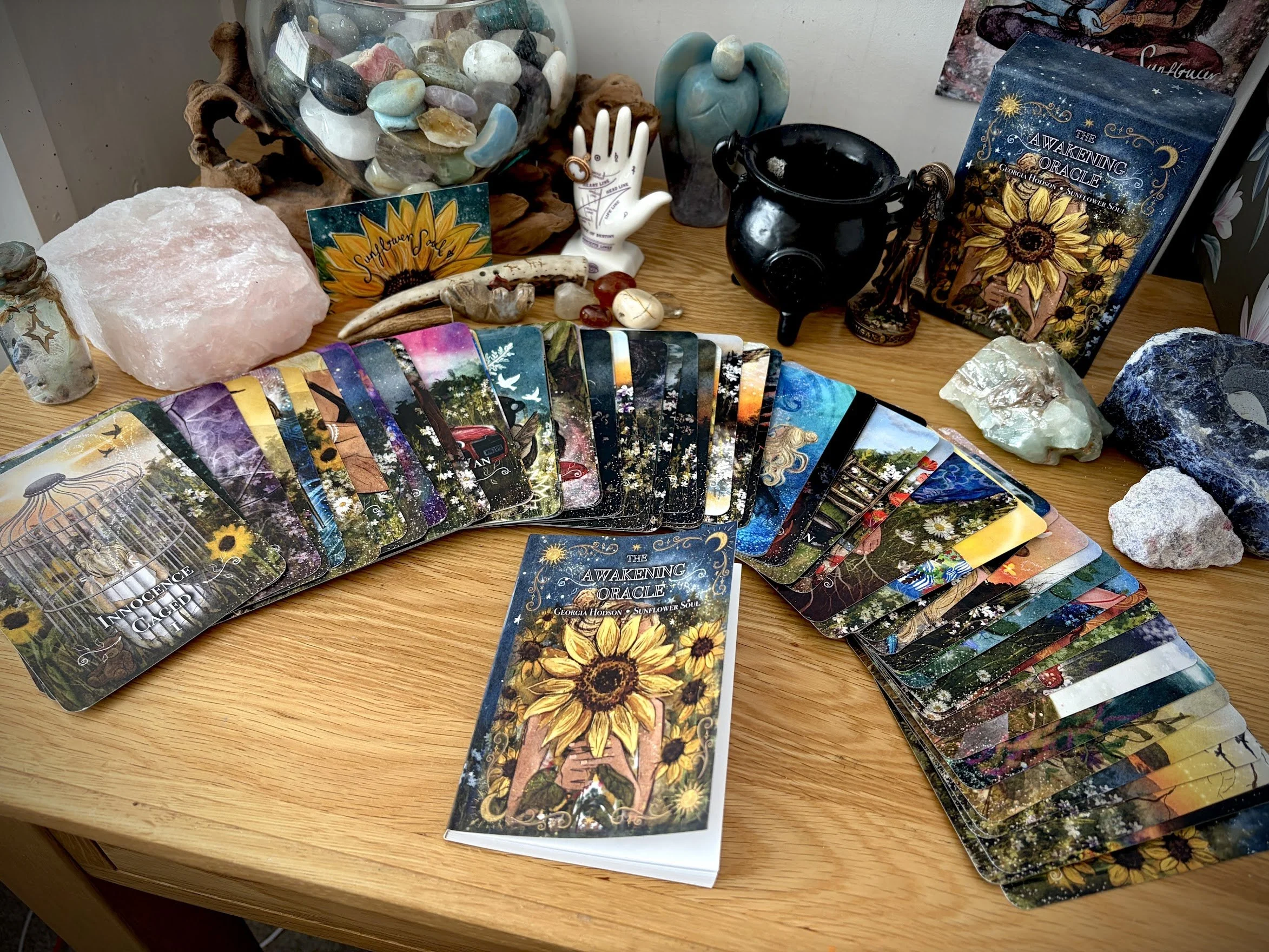 A wooden table displaying tarot cards, a Himalayan salt lamp, quartz crystals, a black cauldron, and various stones and minerals. There is also a book titled 'The Awakening Oracle' and a card with a sunflower illustration that says 'Sunflower Love'.