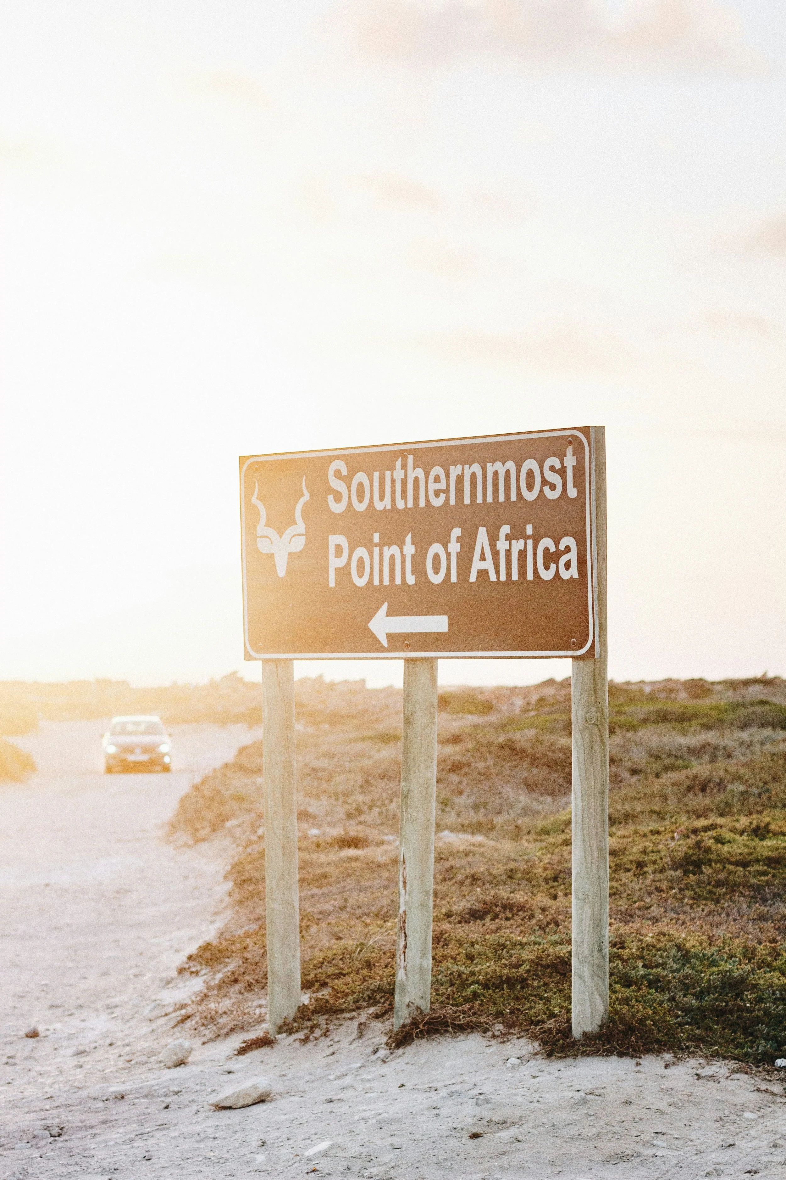 Roadside sign pointing left that reads 'Southernmost Point of Africa' with a decorative logo, in a semi-arid landscape during sunset.