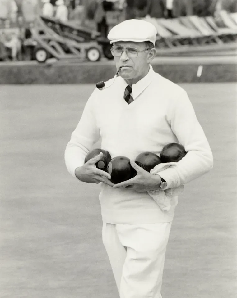 A man dressed in white, wearing a cap and glasses, holding several crown bowling balls on a bowling alley or lane.