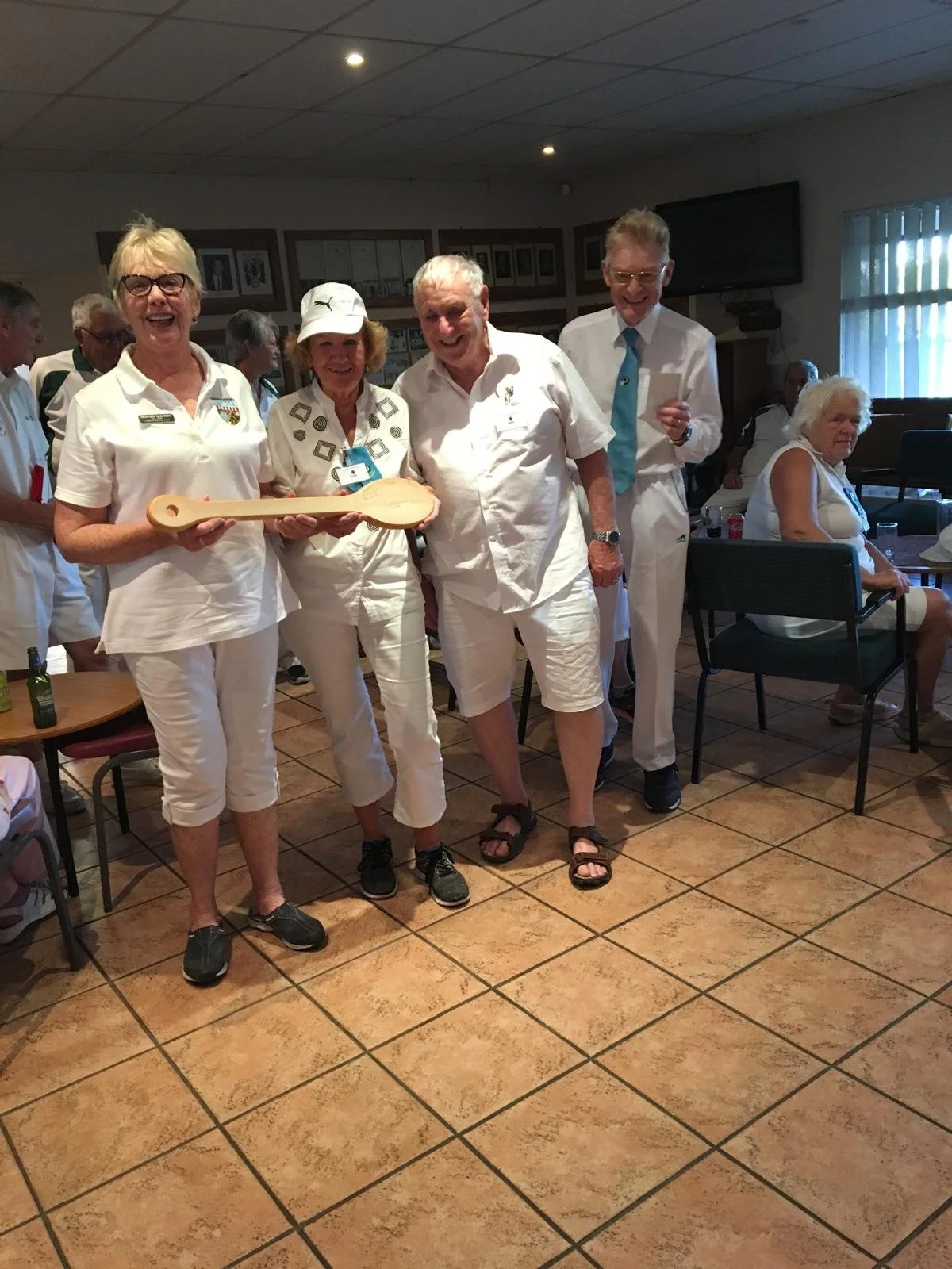 Group of five people in white clothing standing indoors, holding a large wooden spoon, smiling, with other people in the background.