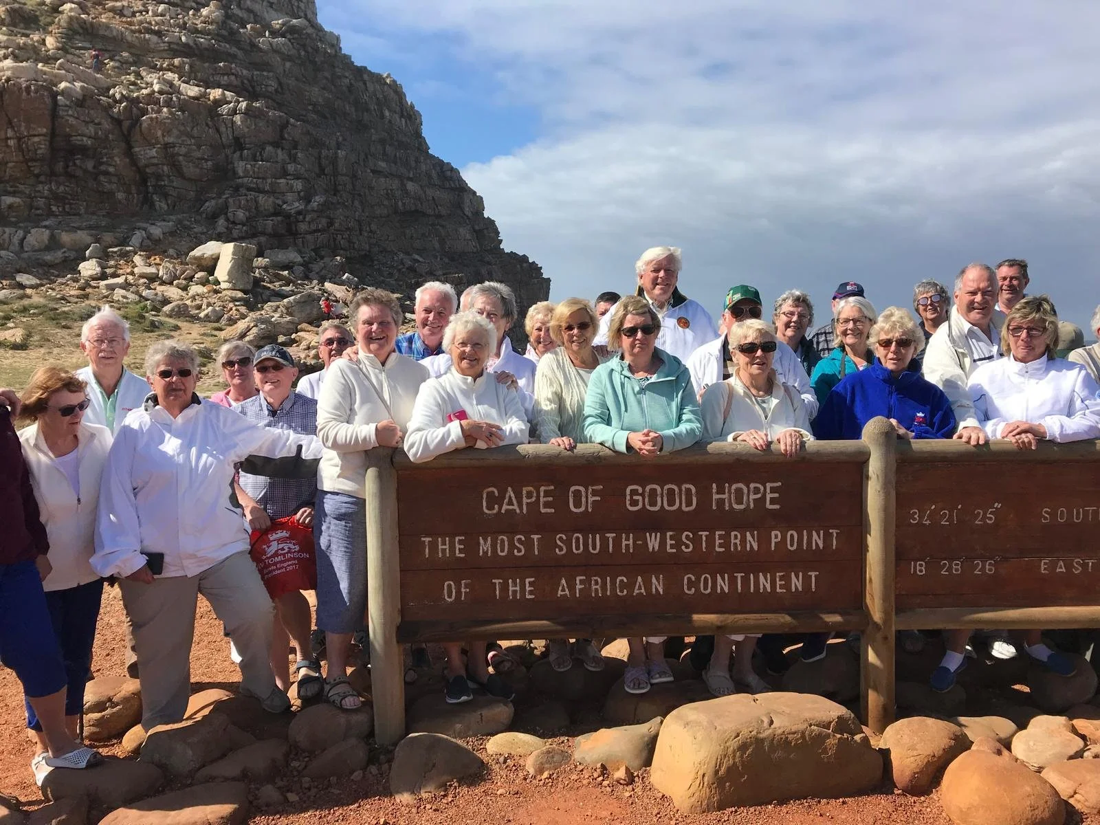 A group of smiling tourists gathered around a sign at Cape of Good Hope, the most south-western point of Africa, with a rocky hillside and partly cloudy sky in the background.