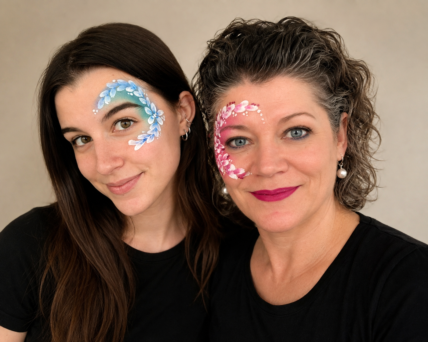 Zwei Frauen mit buntem Gesichtsschmuck in Blau und Pink, lächelnd vor neutralem Hintergrund.