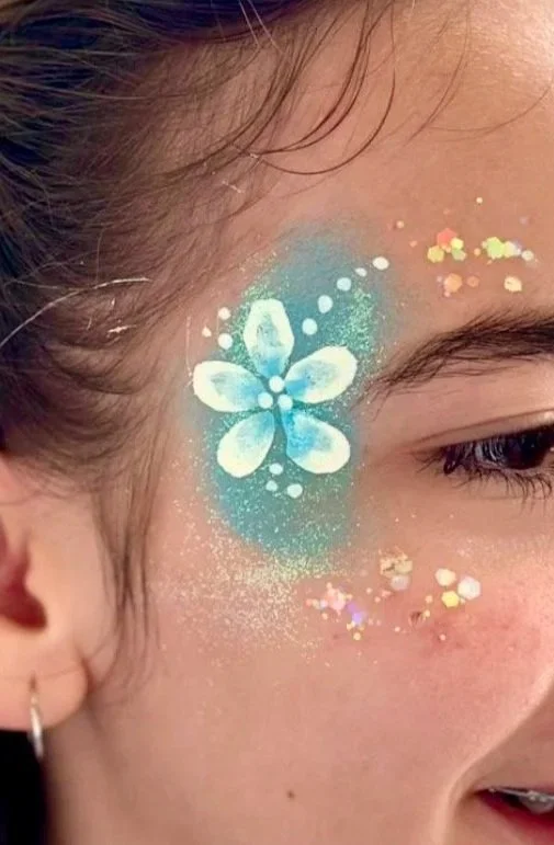 Nahaufnahme eines Gesichts mit buntem, glänzendem Gesichtsschmuck, einschließlich eines blau-weiß leuchtenden Schmetterlings und Glitzersternchen entlang der Wange.