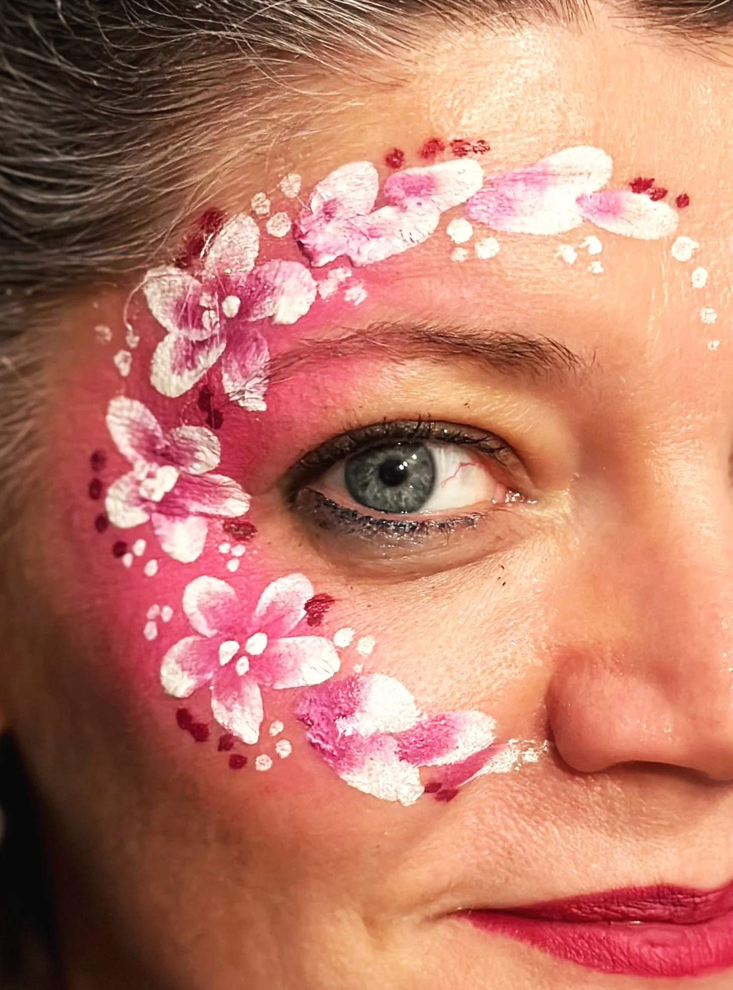 Nahaufnahme eines Gesichts mit Blumen- und Farbschmuck um das Auge, Lippen mit rotem Lippenstift, Haut ist gut beleuchtet.