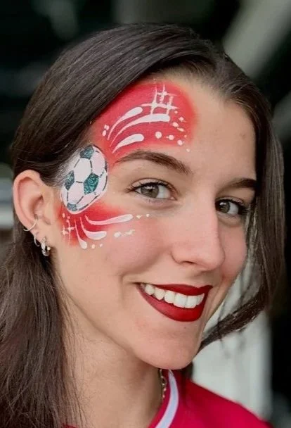 Junge Frau mit sportlichem Gesichtsausdruck, rotem Lippenstift, und Fußball-Tattoo auf der Stirn, trägt rotes Sporttrikot.