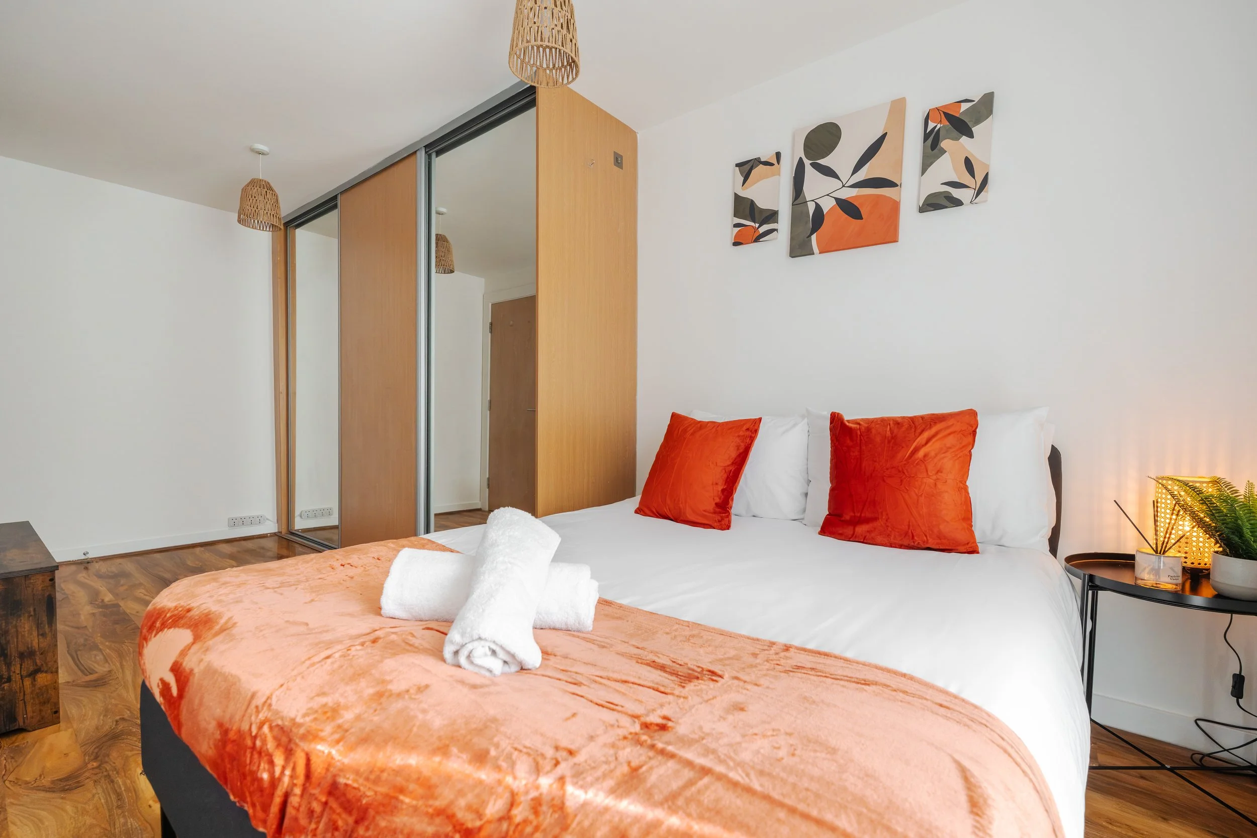 A bedroom with white walls, a wooden closet with sliding mirrored doors, an orange and white bed with orange pillows, a small black side table with a lamp, a potted plant, and wall art featuring abstract autumn-colored leaves.