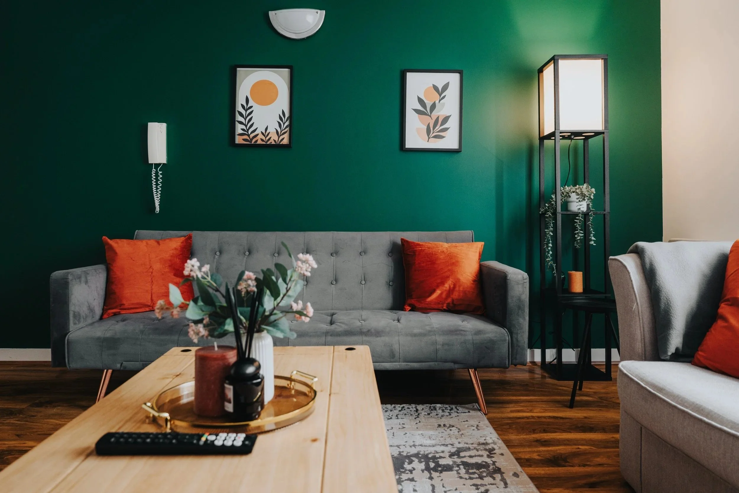 Living room with a gray tufted sofa decorated with orange pillows, a wooden coffee table with a flower vase, a wall with framed artwork and a wall-mounted telephone, and a tall black shelf with a white lamp and a plant.